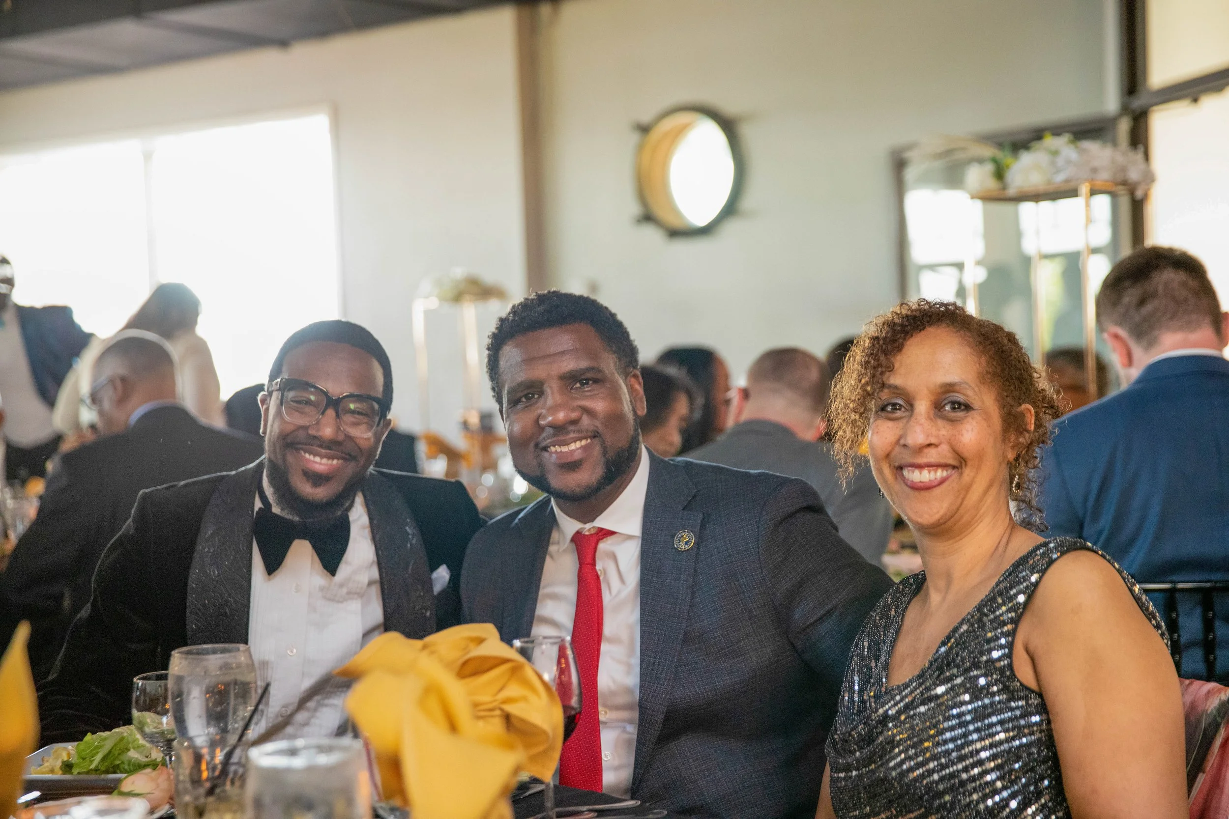 Three people smiling at a formal dinner event with glasses, plates, and decorative yellow napkins on the table.