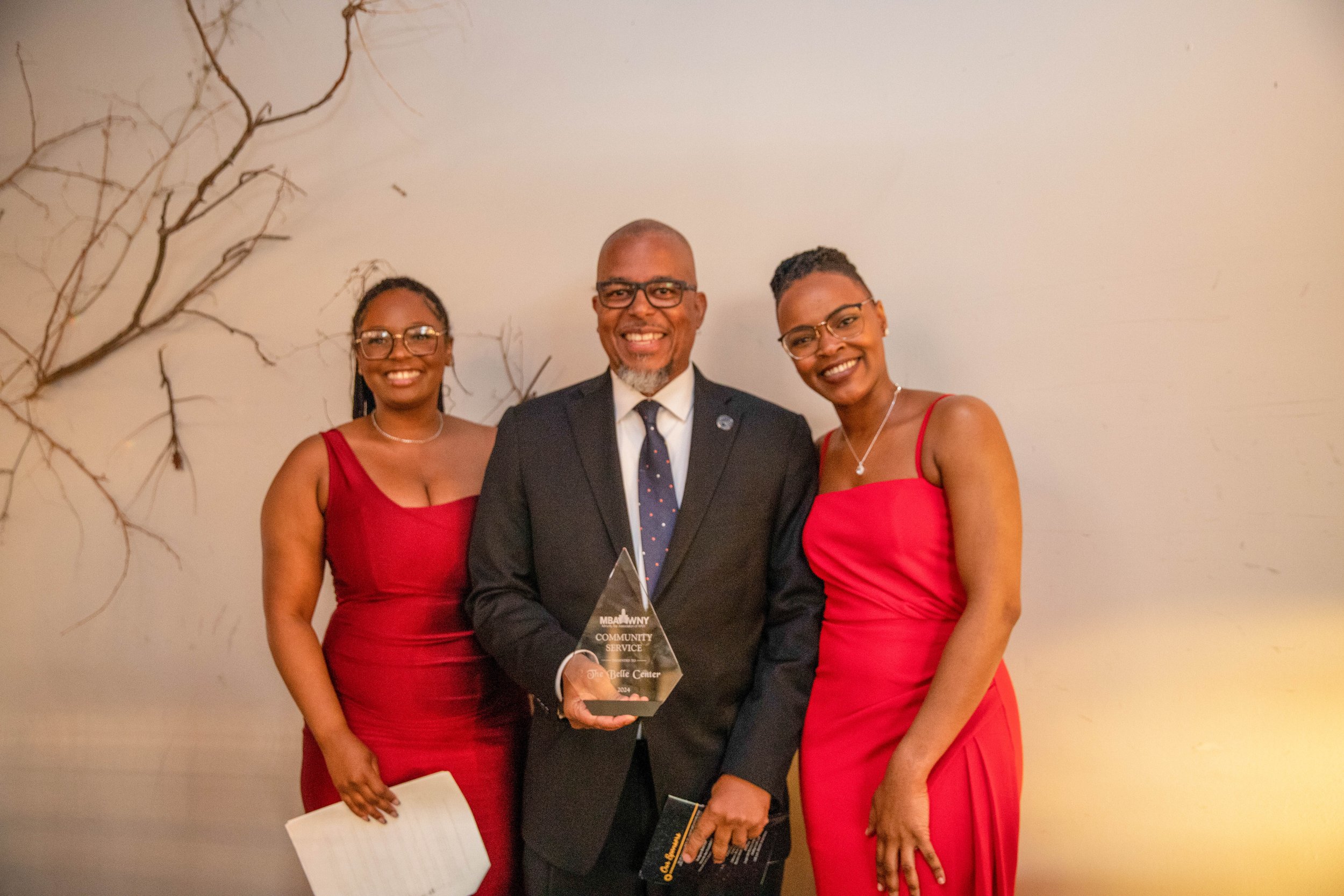 A group of three people, two women and one man, standing together in formal attire. The man is in the center holding a clear triangular award with text on it. The women are wearing red dresses, and all three are smiling. They are standing against a p