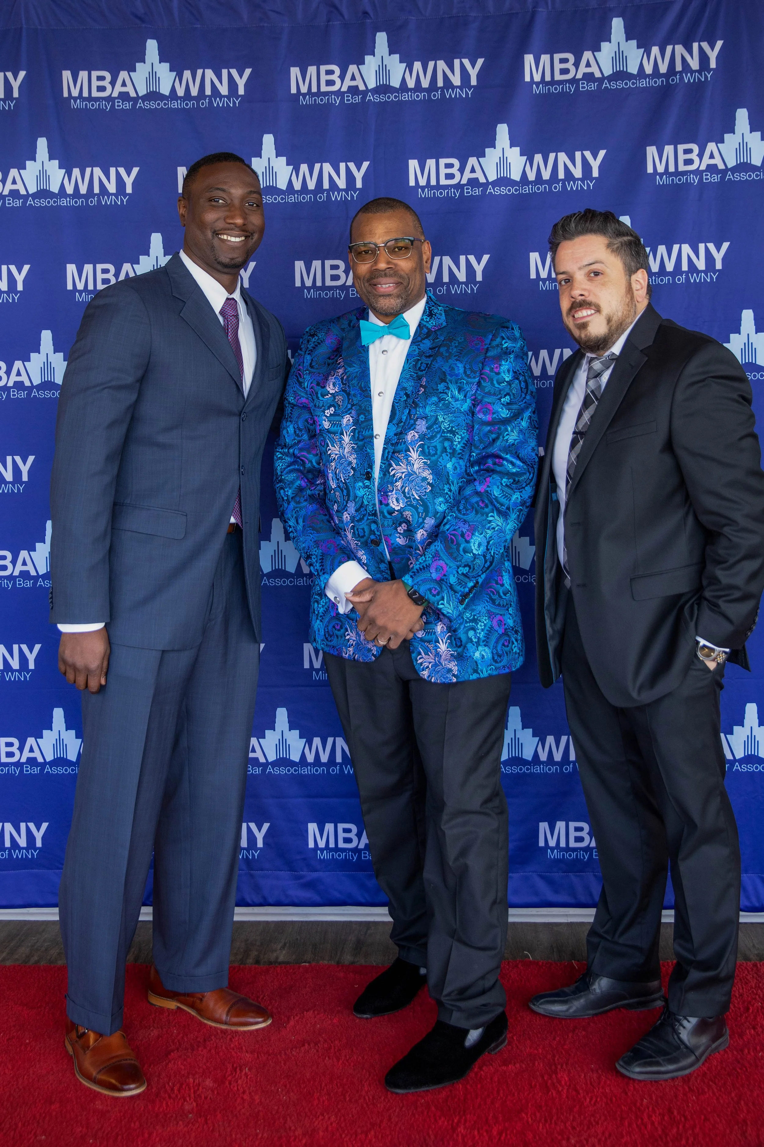 Three men dressed in formal suits standing on a red carpet in front of a blue backdrop with the logo of the Minority Bar Association of WNY. The man in the middle is wearing a bright blue patterned suit with a matching bow tie, while the other two me