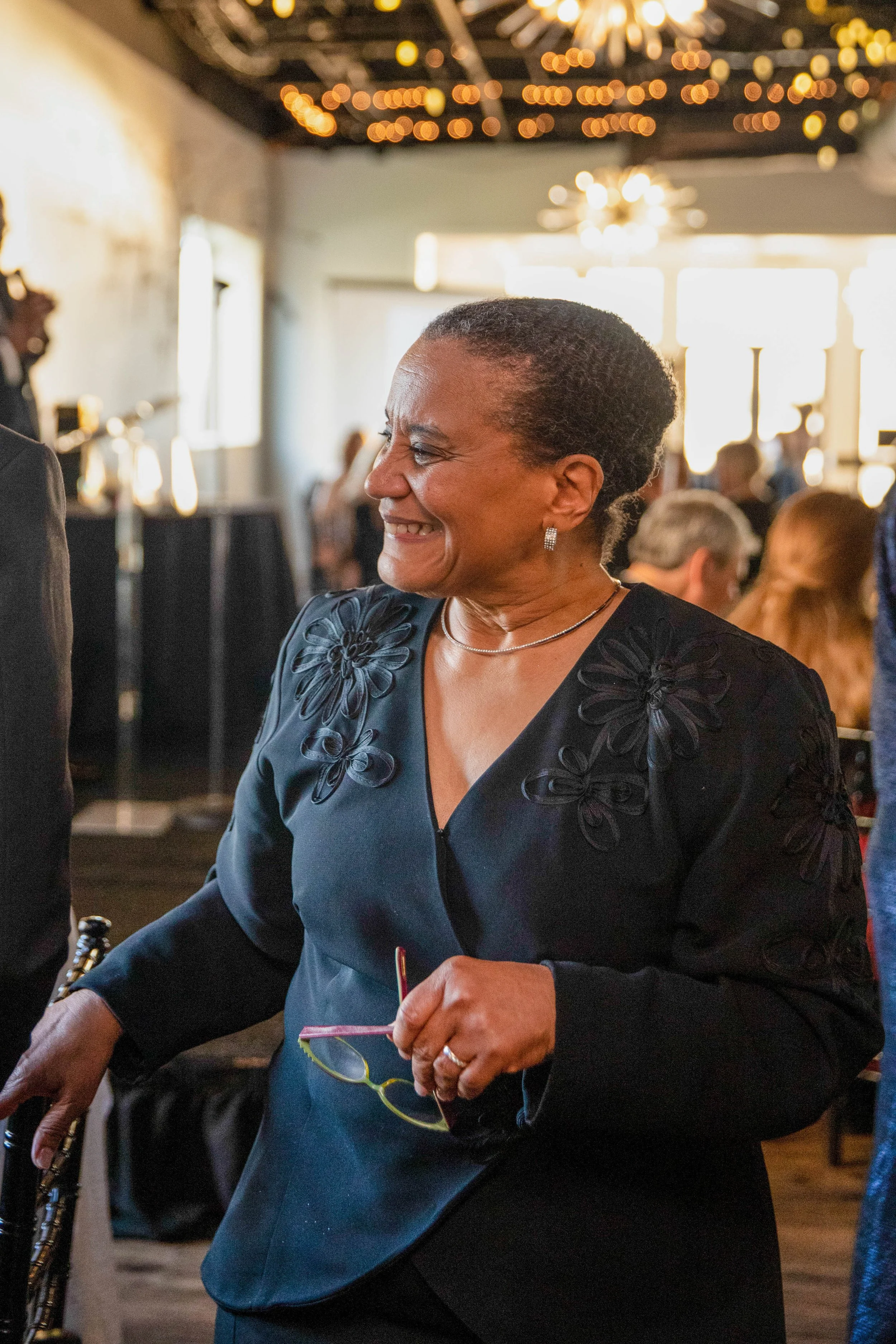 A smiling woman wearing a black dress with floral embroidery, earrings, and a necklace, holding a knitting project with multicolored yarn at an indoor event or gathering.