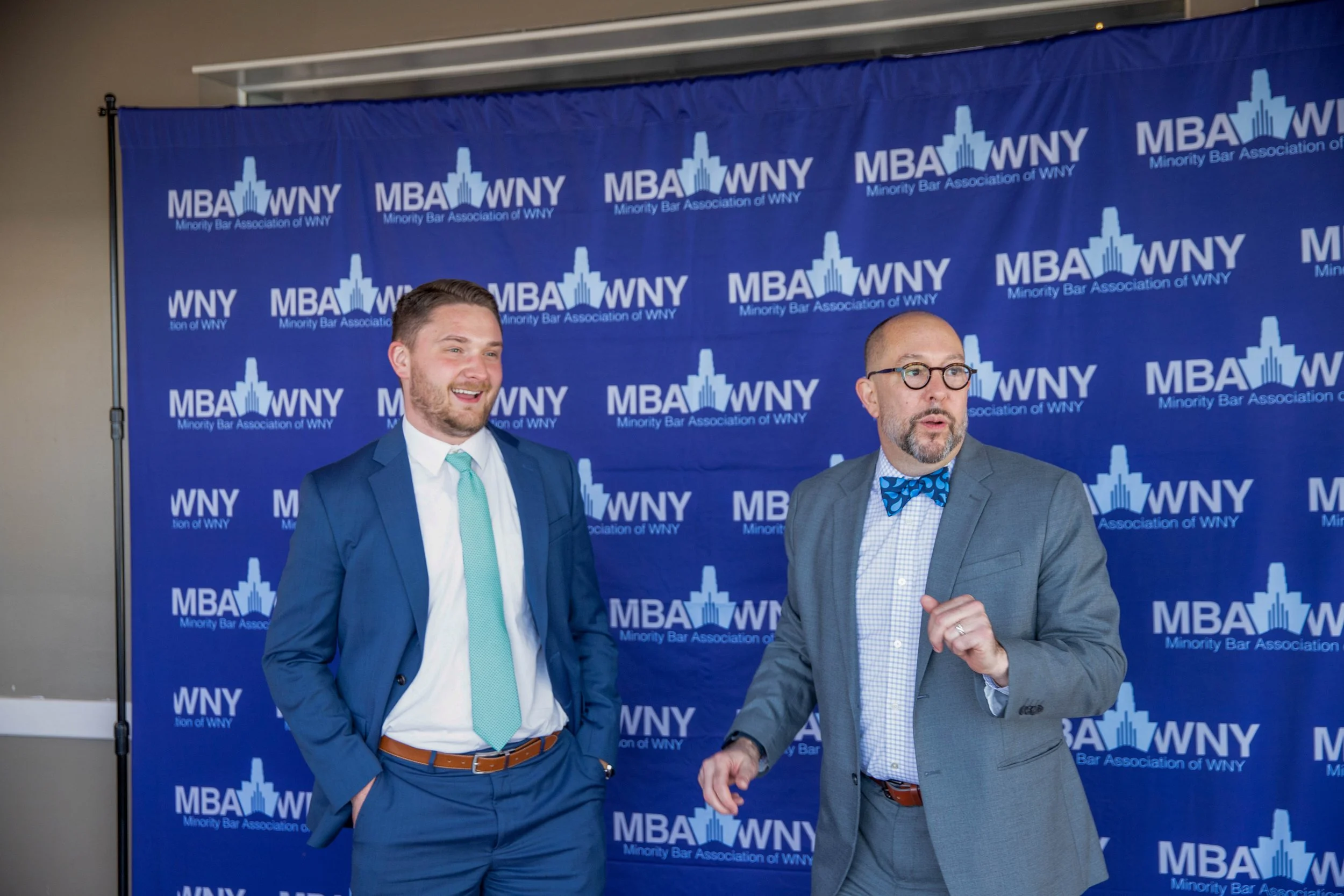 Two men in suits standing in front of a blue backdrop with the logo and text 'MBA WNY Minority Bar Association of WNY'. One man has a blue suit, teal tie, and brown belt, smiling. The other man, with glasses and a beard, wears a gray suit with a blue