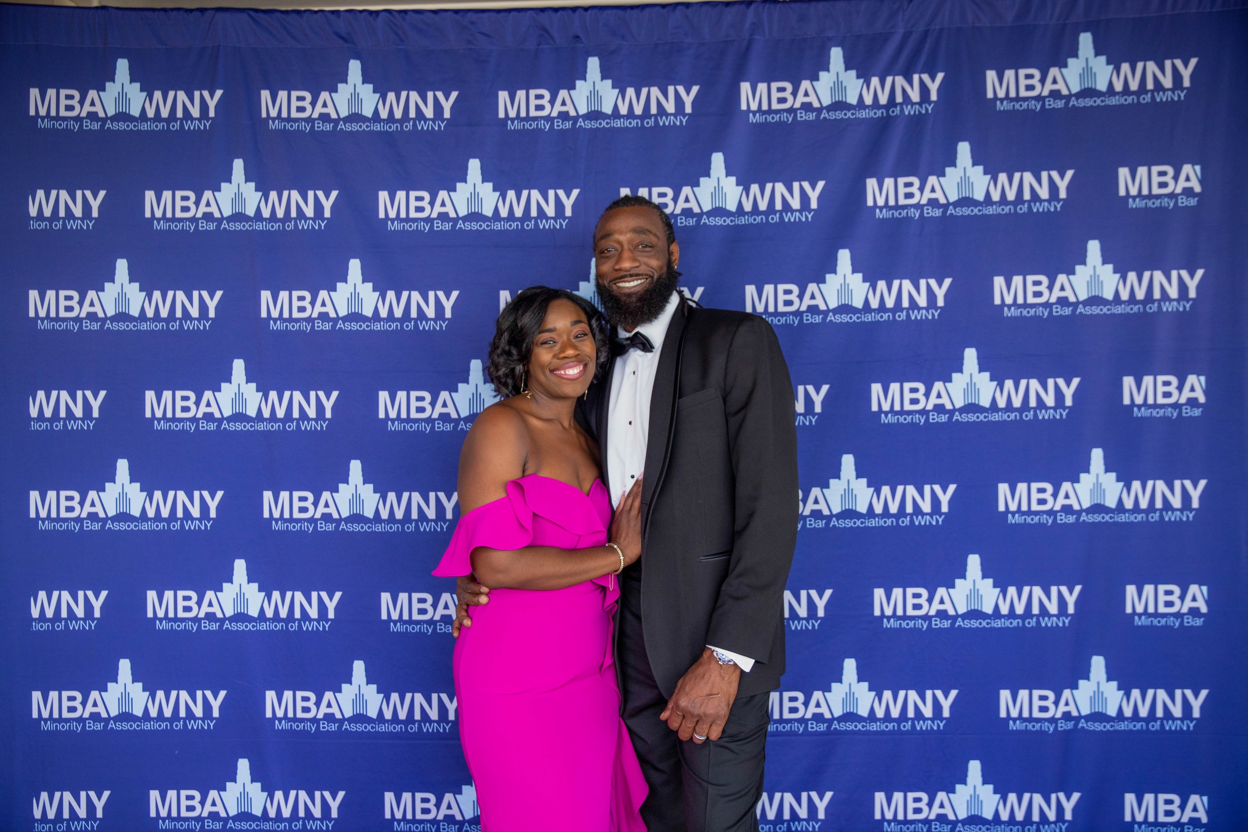 A man and woman dressed in formal attire are standing together, smiling, against a blue backdrop with the text 'MBA WNY Minority Bar Association of WNY' and a city skyline logo repeated across it.