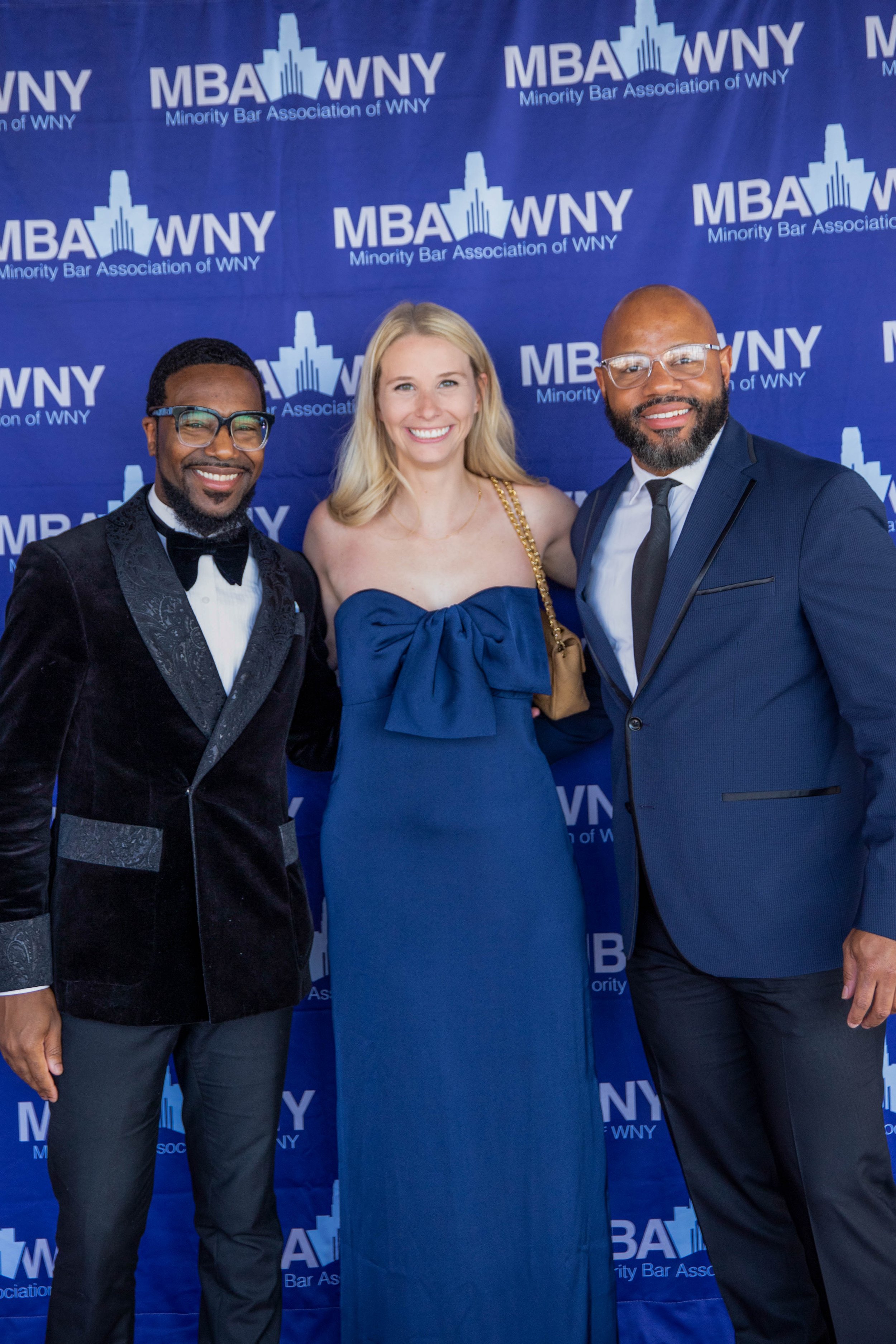 Three people standing in front of a blue backdrop with the text 'Minority Bar Association of WNY' and 'MBA WNY' logo. They are dressed formally, smiling, and looking at the camera.