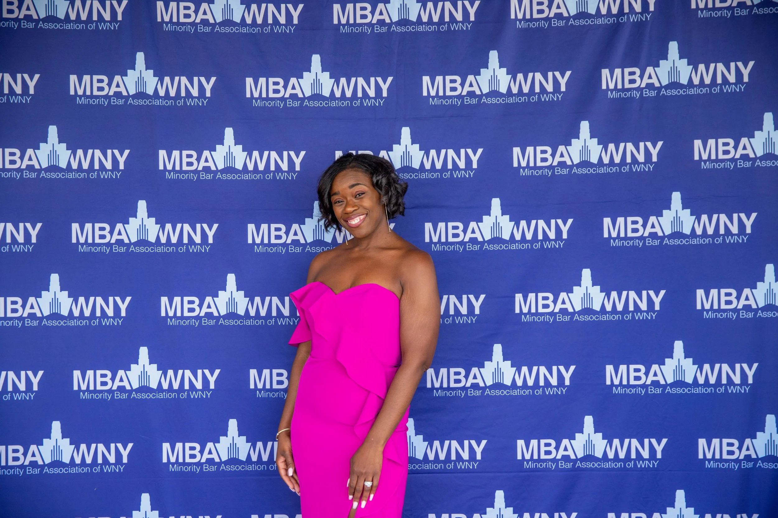 A woman in a bright pink dress standing in front of a blue backdrop with the logo of the Minority Bar Association of WNY, smiling at the camera.