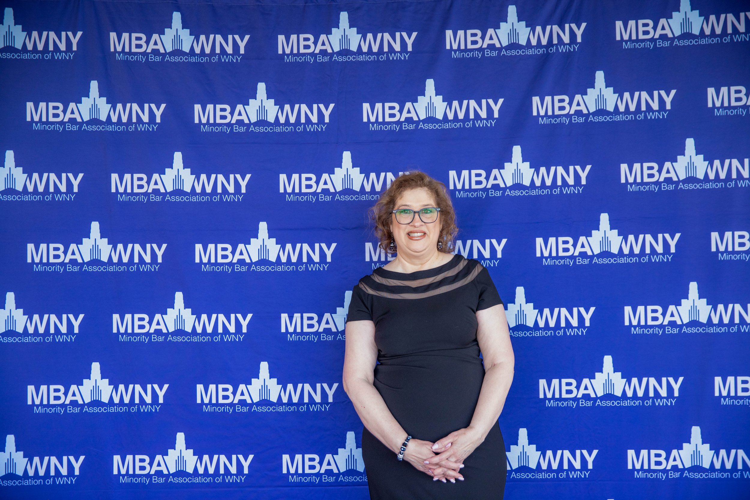 A woman with glasses, curly hair, and a black dress standing in front of a blue backdrop with the logo of the Minority Bar Association of WNY.