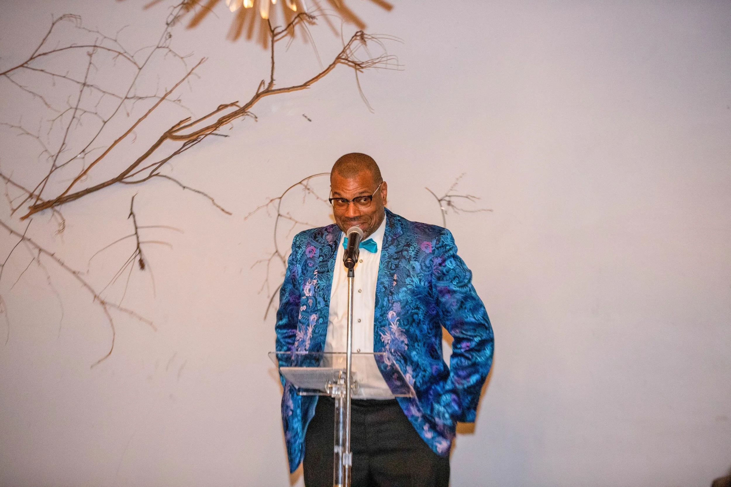 A man wearing glasses and a brightly colored, patterned blazer with a bow tie is speaking into a microphone at a podium, with a white wall and decorative branches in the background.