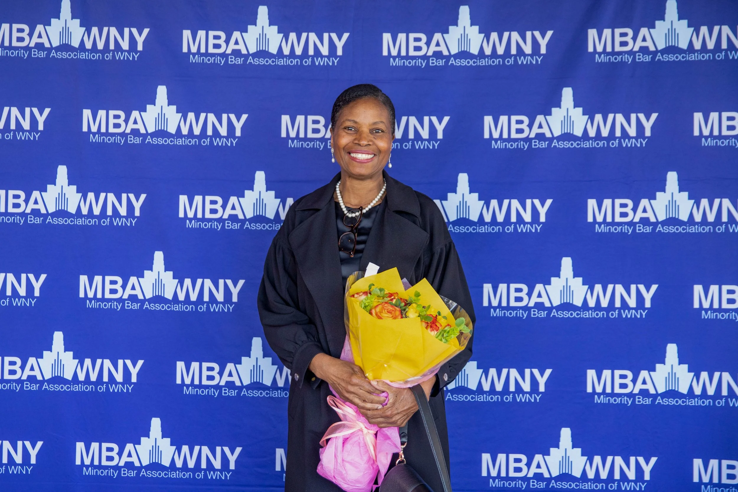 A smiling woman holding a bouquet of flowers stands in front of a blue backdrop with the logo and name of the Minority Bar Association of WNY, MBA WNY.