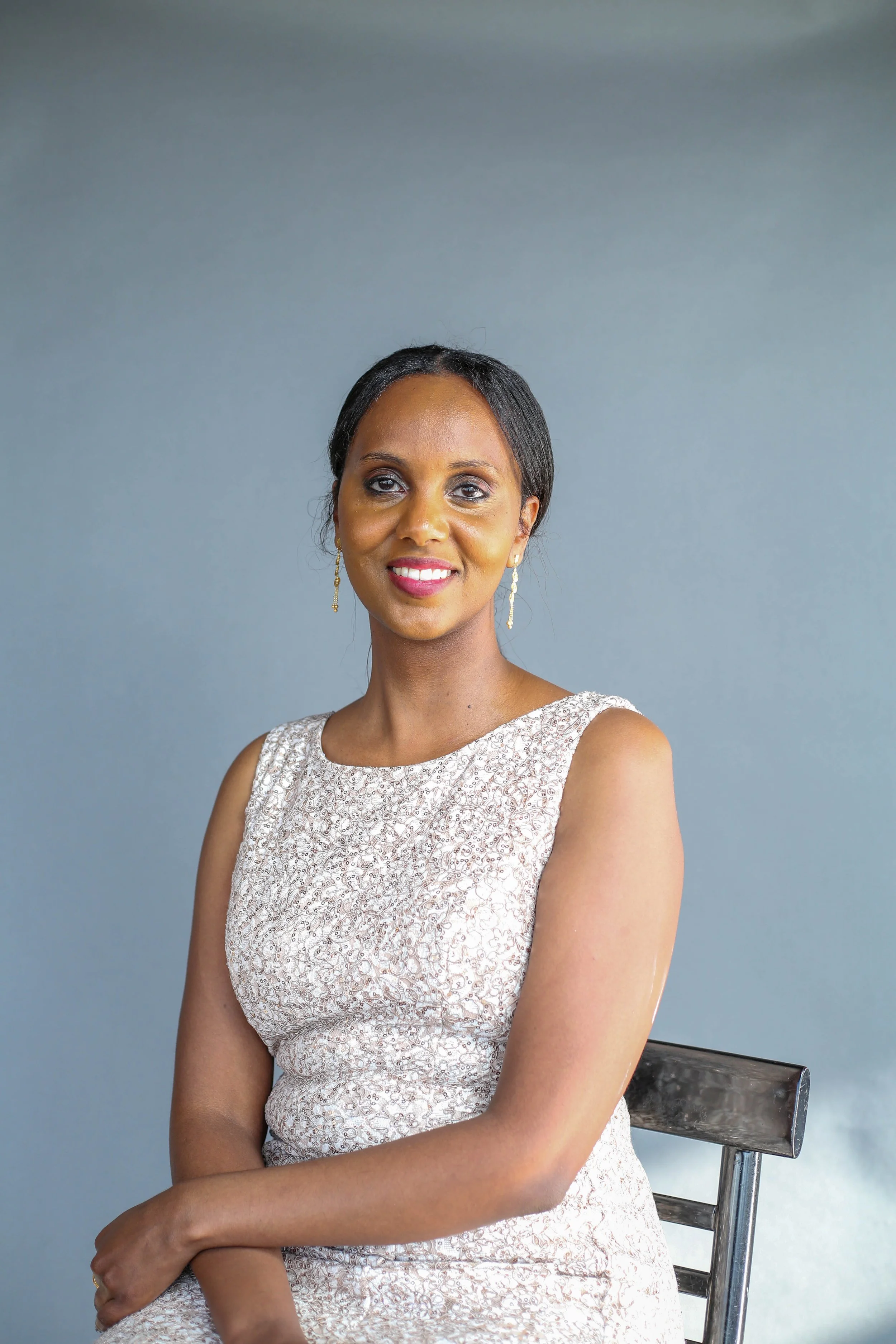 A woman with dark hair and earrings, wearing a sleeveless lace dress, seated on a wooden chair against a plain blue background and smiling.