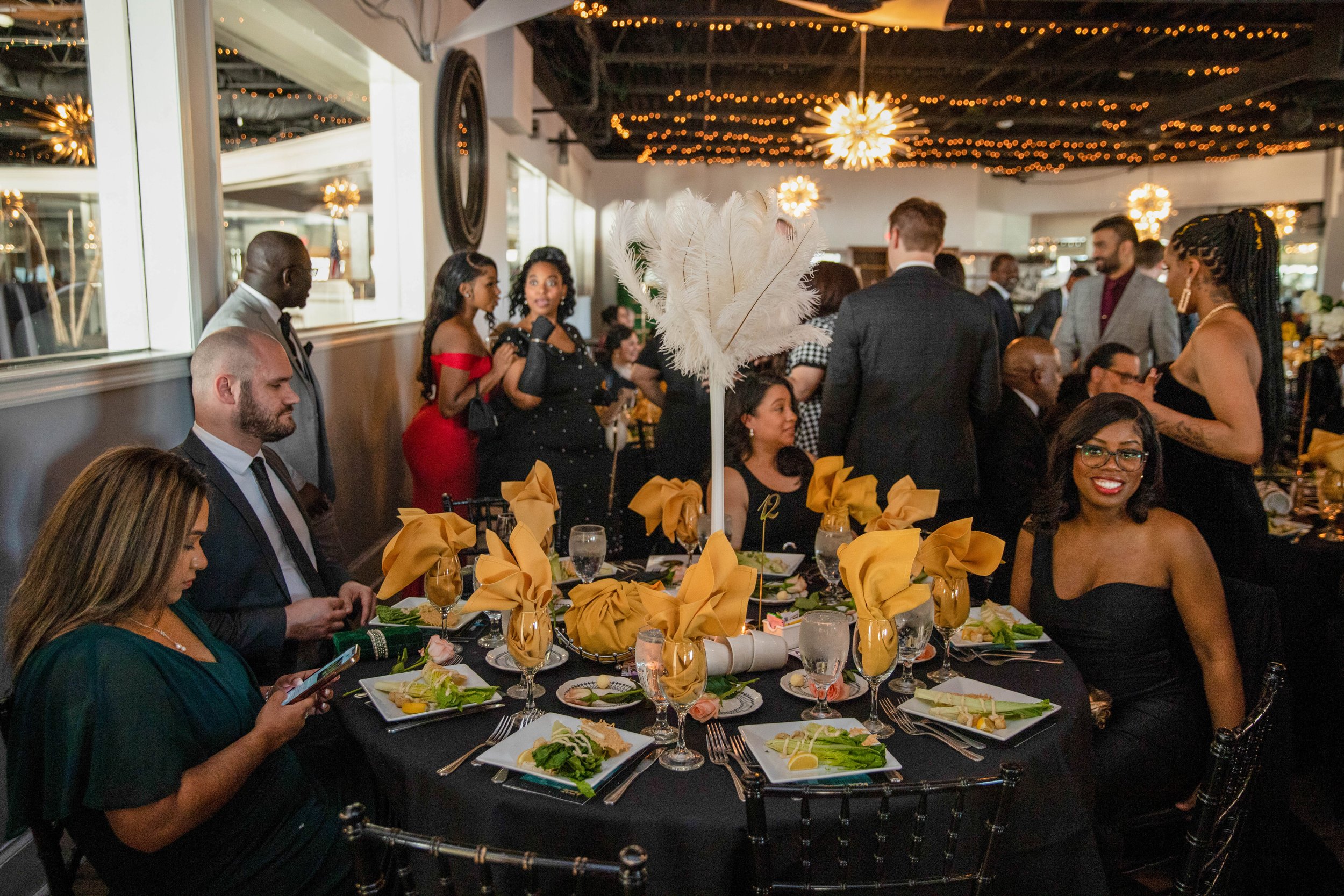 People gathered at a formal dinner event with decorated tables and a fluffy feather centerpiece in a well-lit room with string lights and chandeliers hanging from the ceiling.
