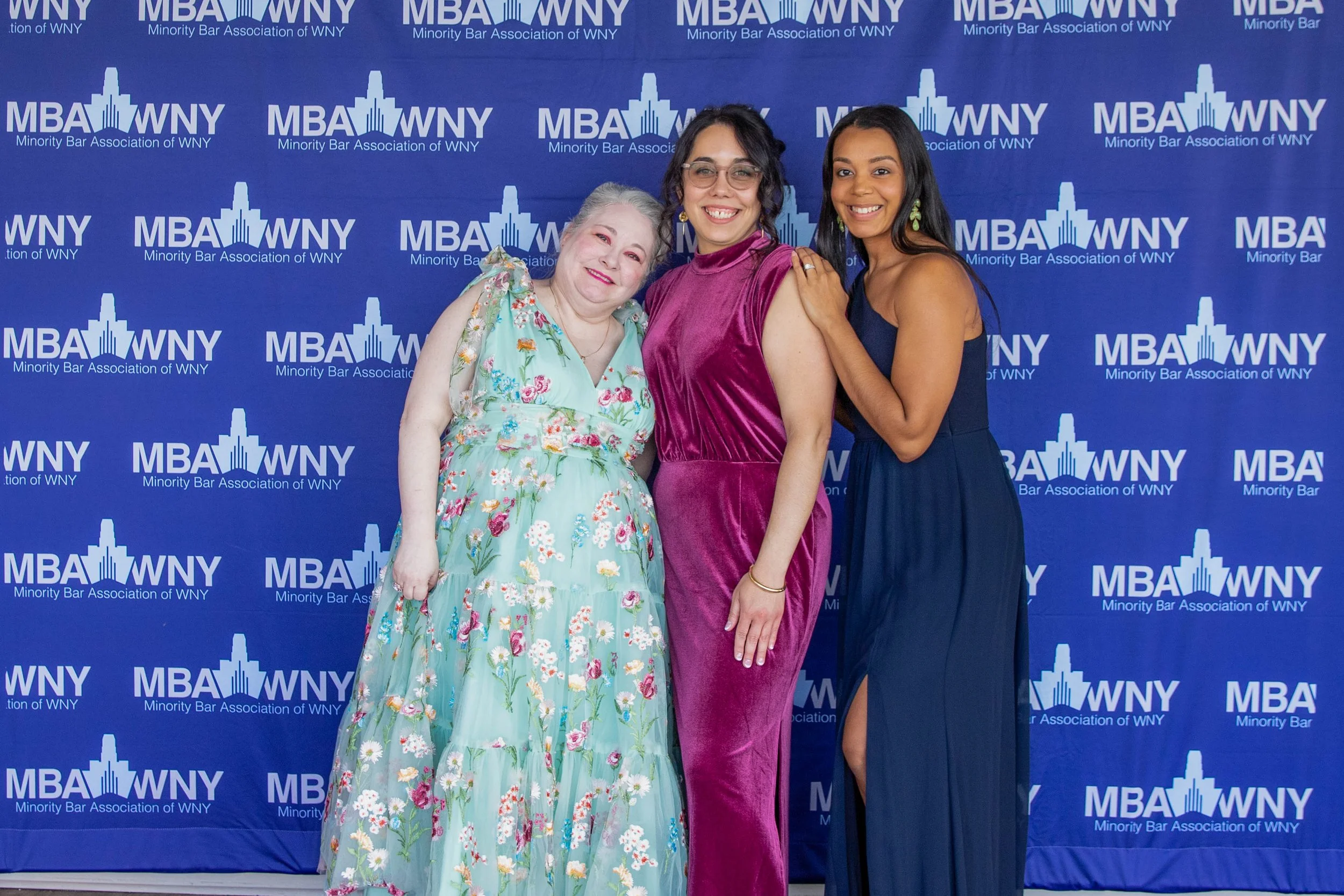 Three women standing in front of a blue backdrop with the logo and name of the Minority Bar Association of WNY. They are smiling and dressed in formal attire. The woman on the left wears a floral dress, the woman in the middle wears a velvety purple 