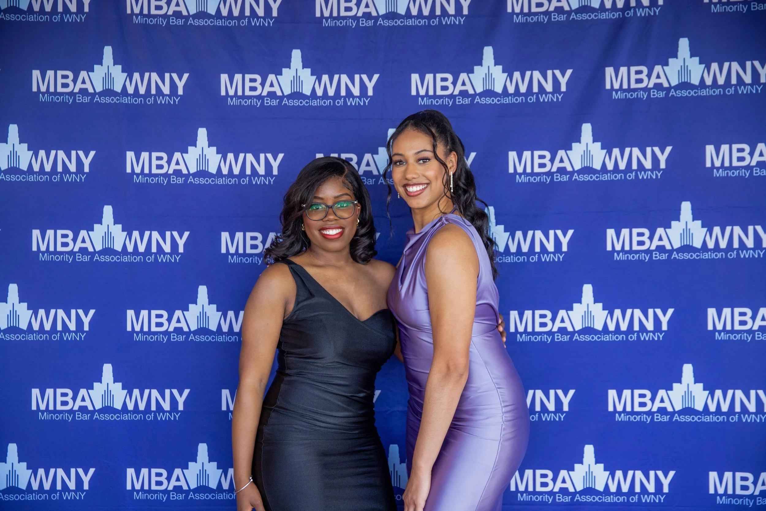 Two women standing in front of a blue backdrop with the text 'MBA WNY Minority Bar Association of WNY' repeated multiple times. Both women are smiling, dressed in elegant evening wear, and leaning slightly toward each other.