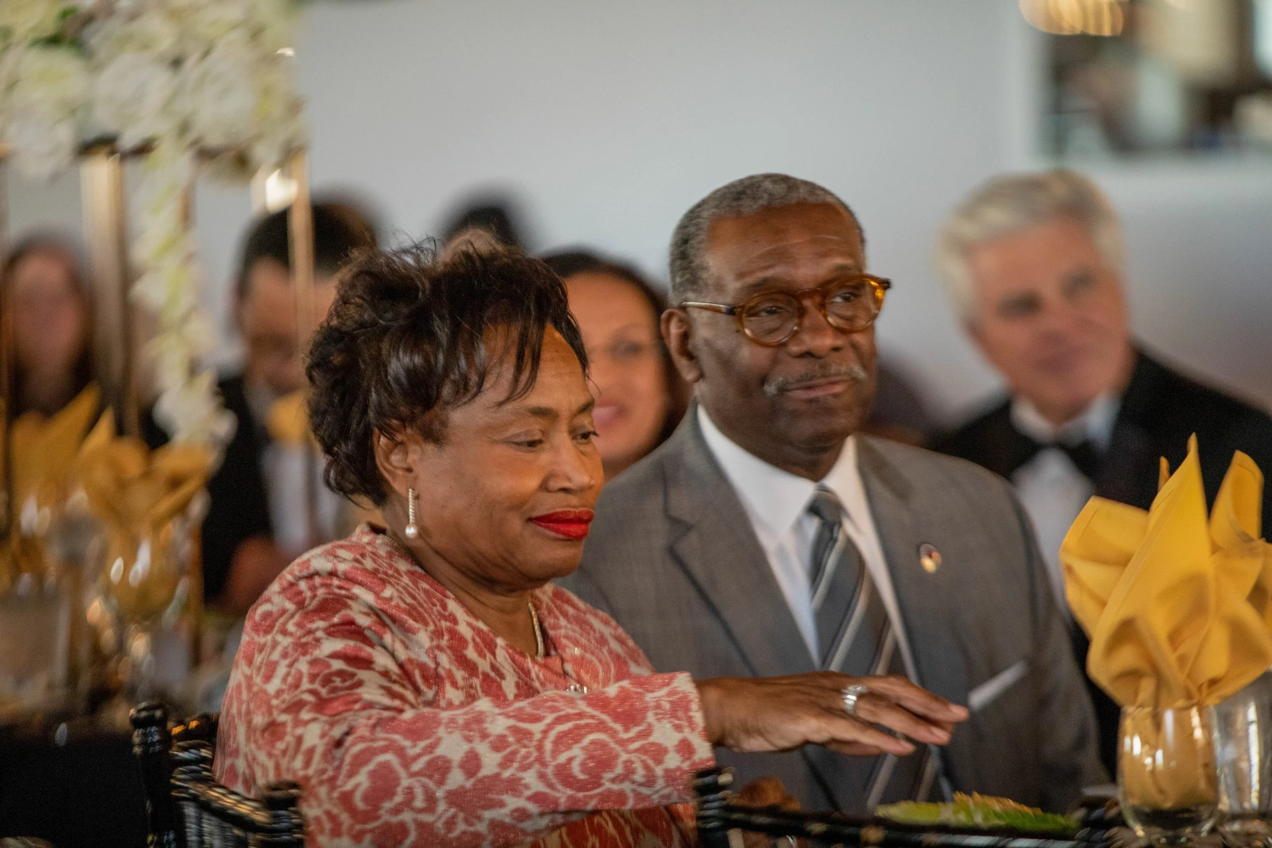 People attending a formal event seated at a table with decorative flowers and yellow napkins.