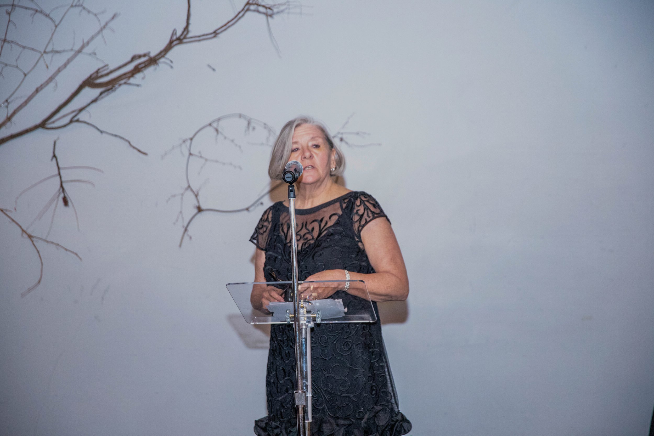 An elderly woman with gray hair, wearing a black lace dress, standing behind a clear podium and speaking into a microphone at an indoor event with a plain light-colored wall background and decorative branches on the side.