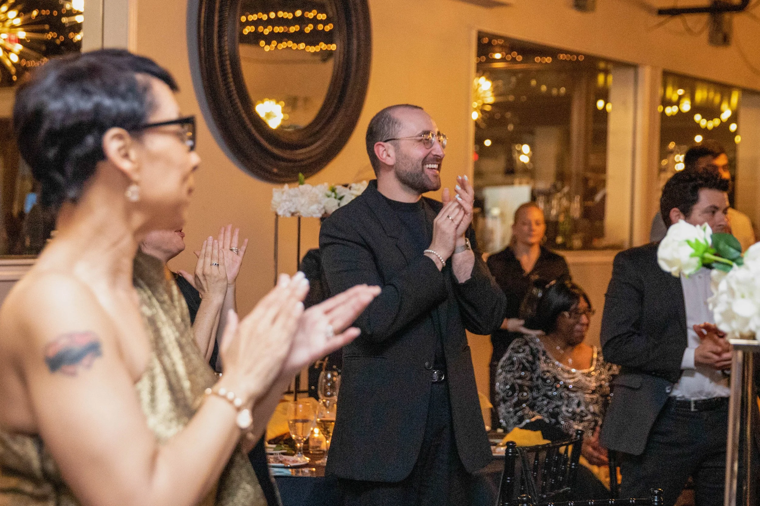 People standing and clapping at an indoor event with warm lighting, including a woman with glasses and a tattoo on her arm, and a man with a beard and glasses smiling. Drinks and floral arrangements are on the tables.