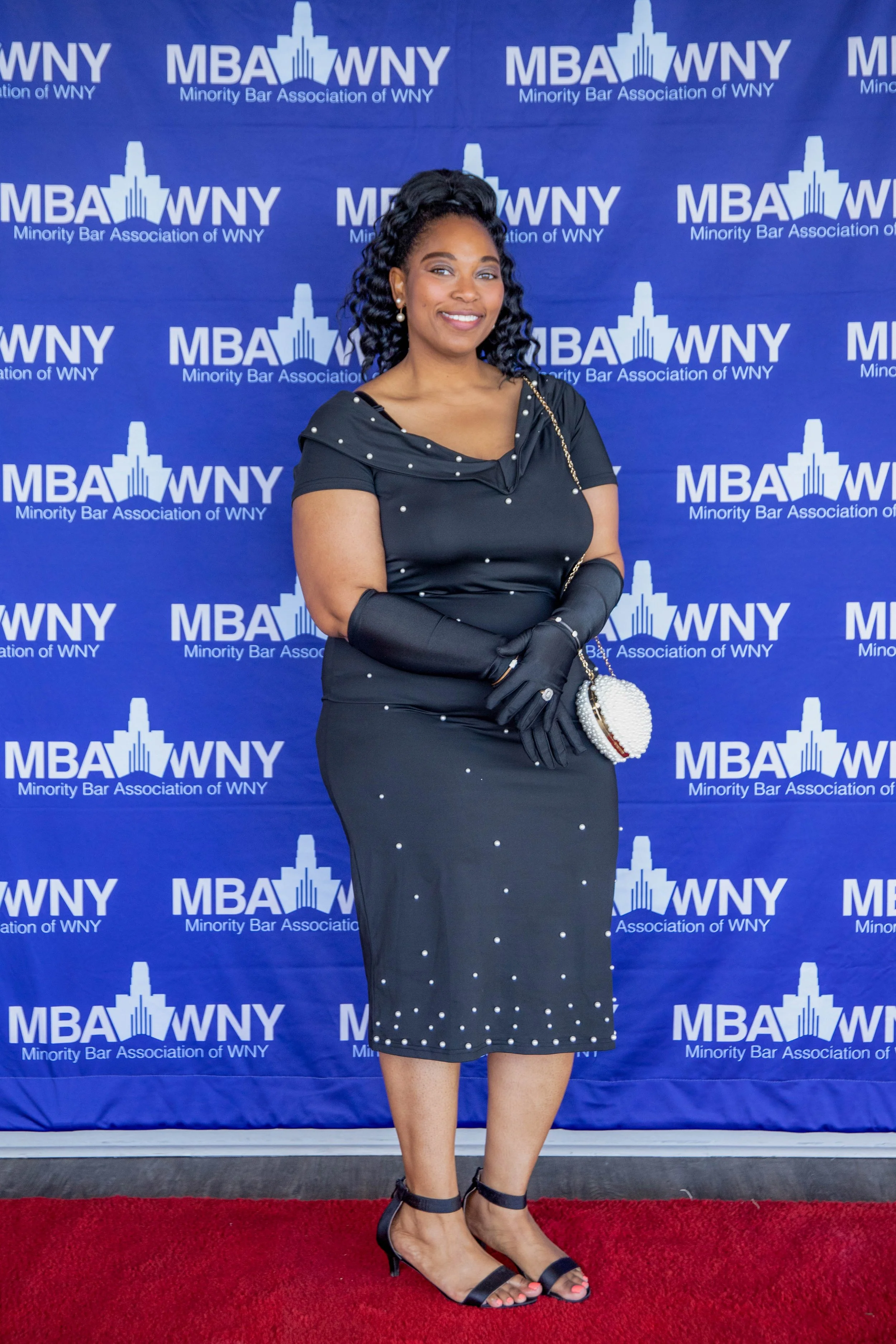 A woman dressed in a black, polka-dot dress and black heels stands on a red carpet in front of a blue backdrop with the logo and name of the Minority Bar Association of WNY. She is smiling, wearing earrings, pearls, and a black glove, holding a pearl