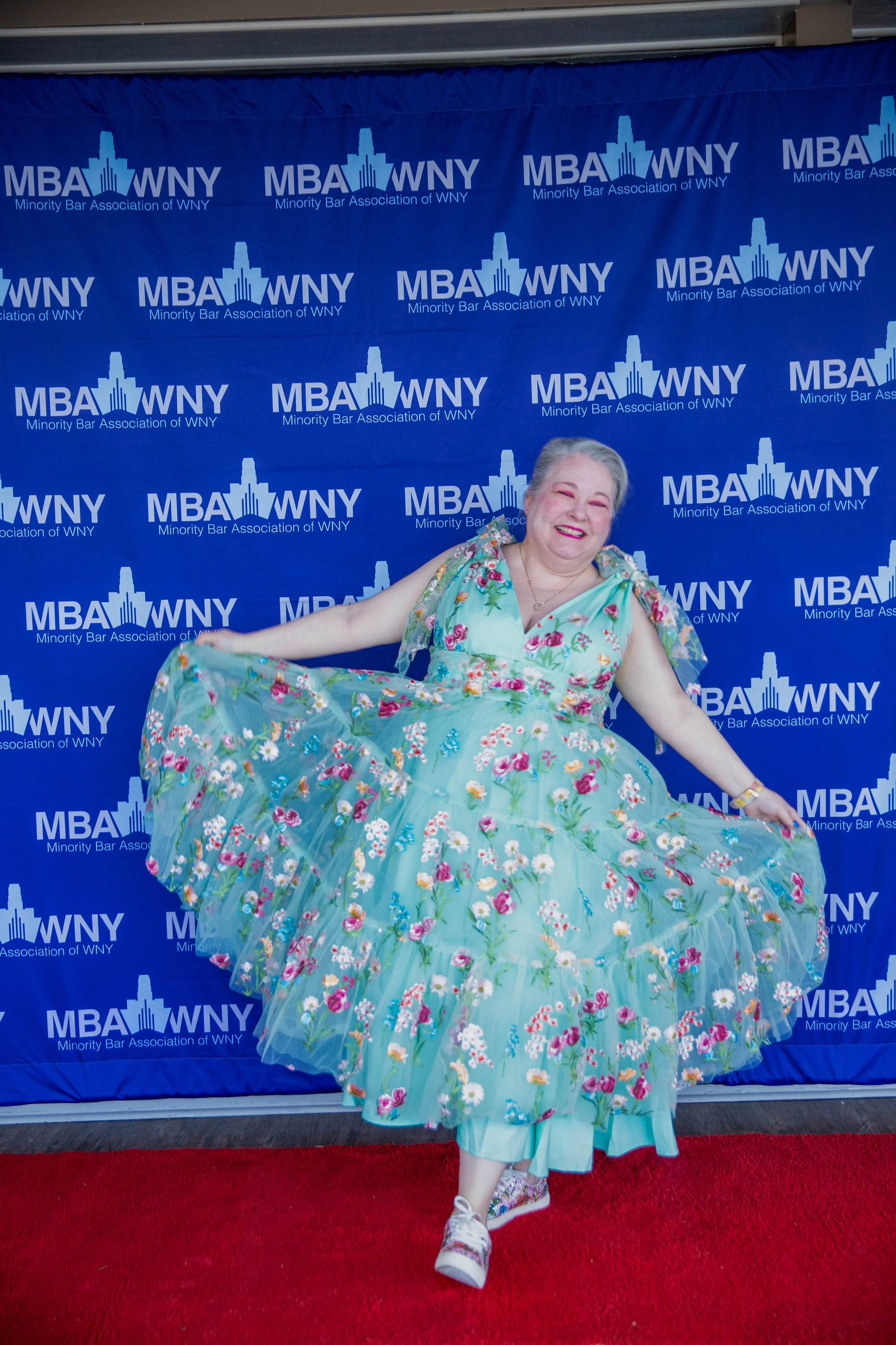 A woman in a floral dress appears to be dancing or performing in front of a blue backdrop with the logo of the Minority Bar Association of WNY repeated multiple times.