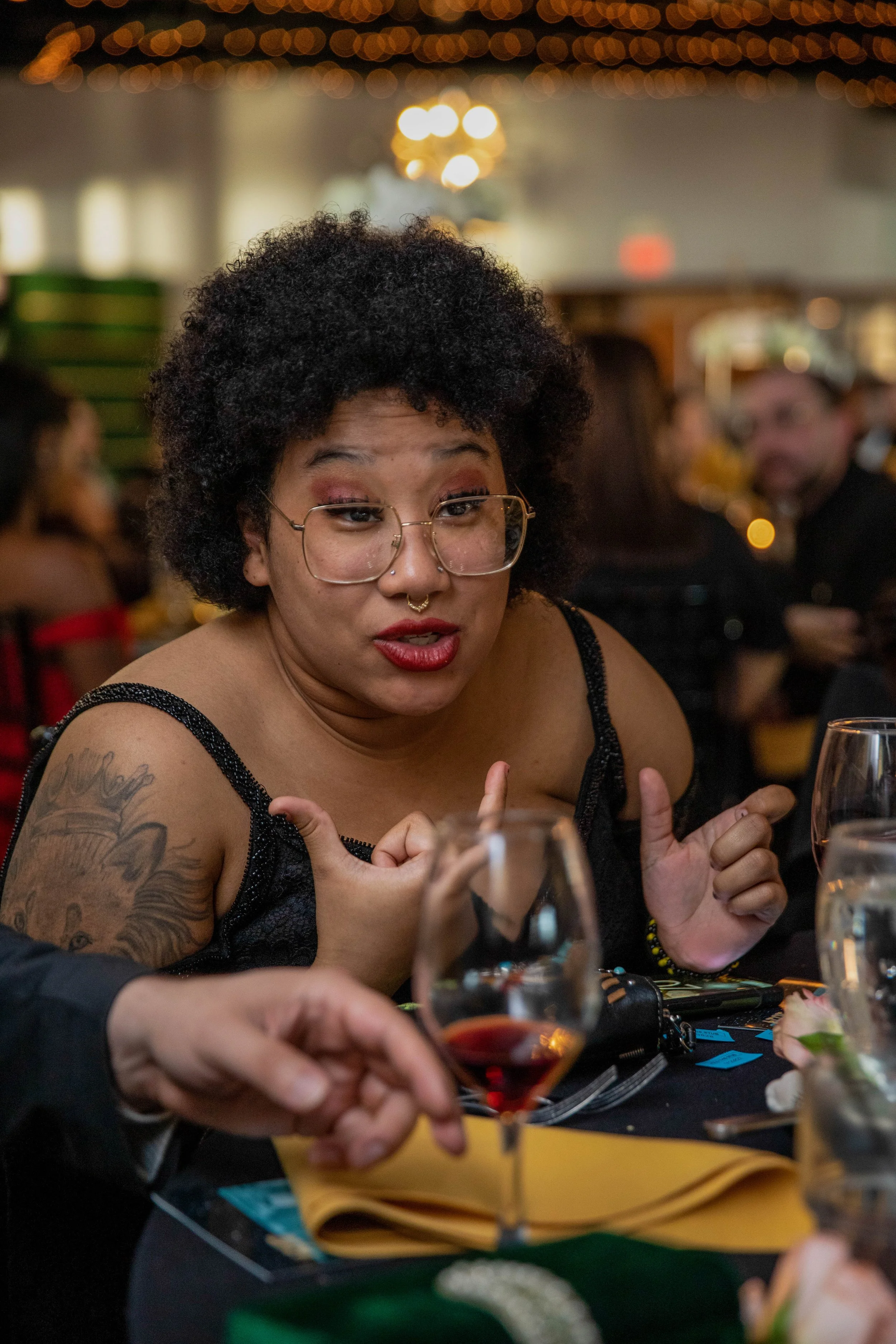 A woman with curly black hair, glasses, and tattoos on her arm, is sitting at a table in a dimly lit restaurant. She appears to be speaking or gesturing with her hands while holding a drink. There are other people and glasses on the table, and the ba