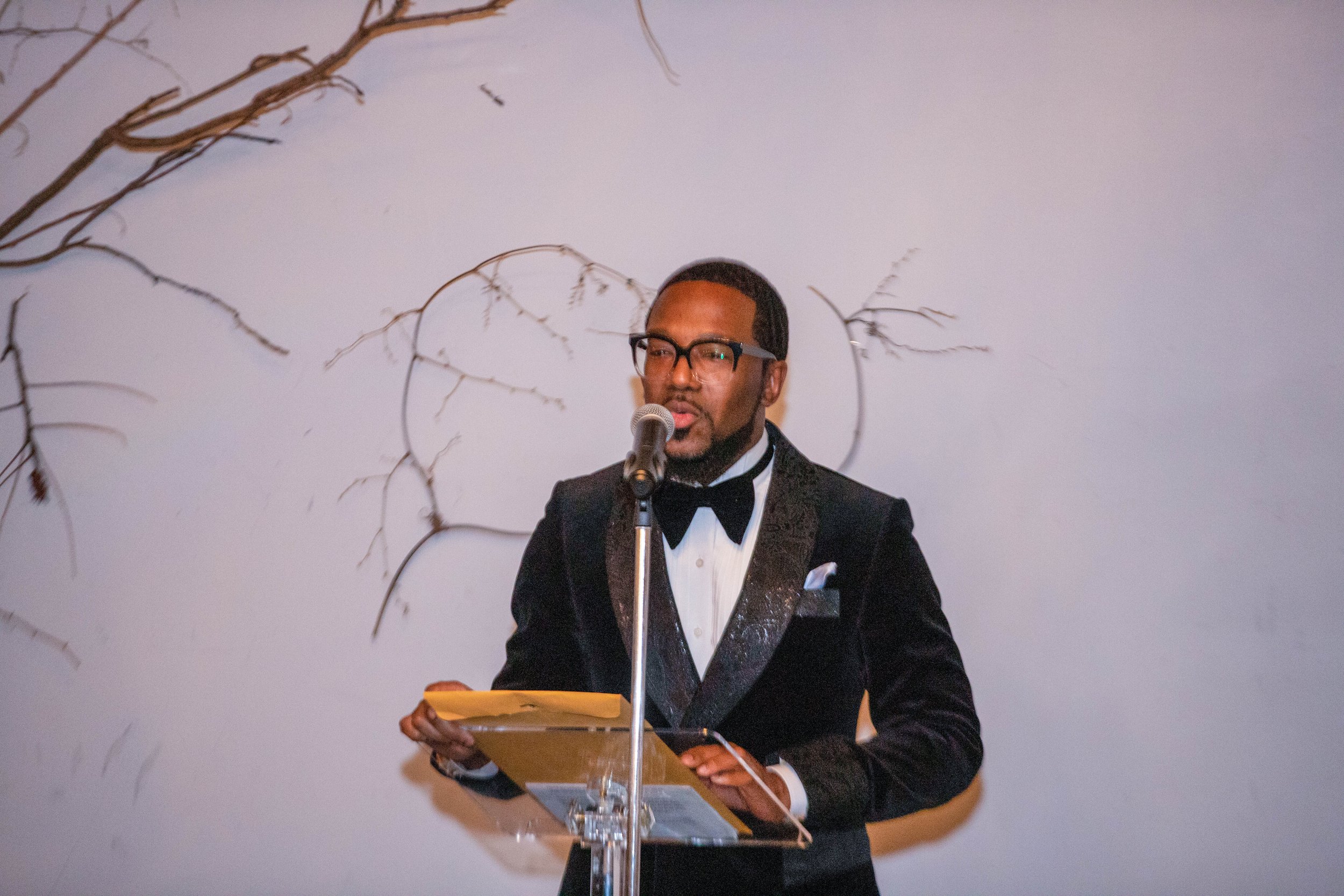 A man dressed in a tuxedo with glasses standing behind a podium, speaking into a microphone, with a plain wall decorated with dried branches in the background.