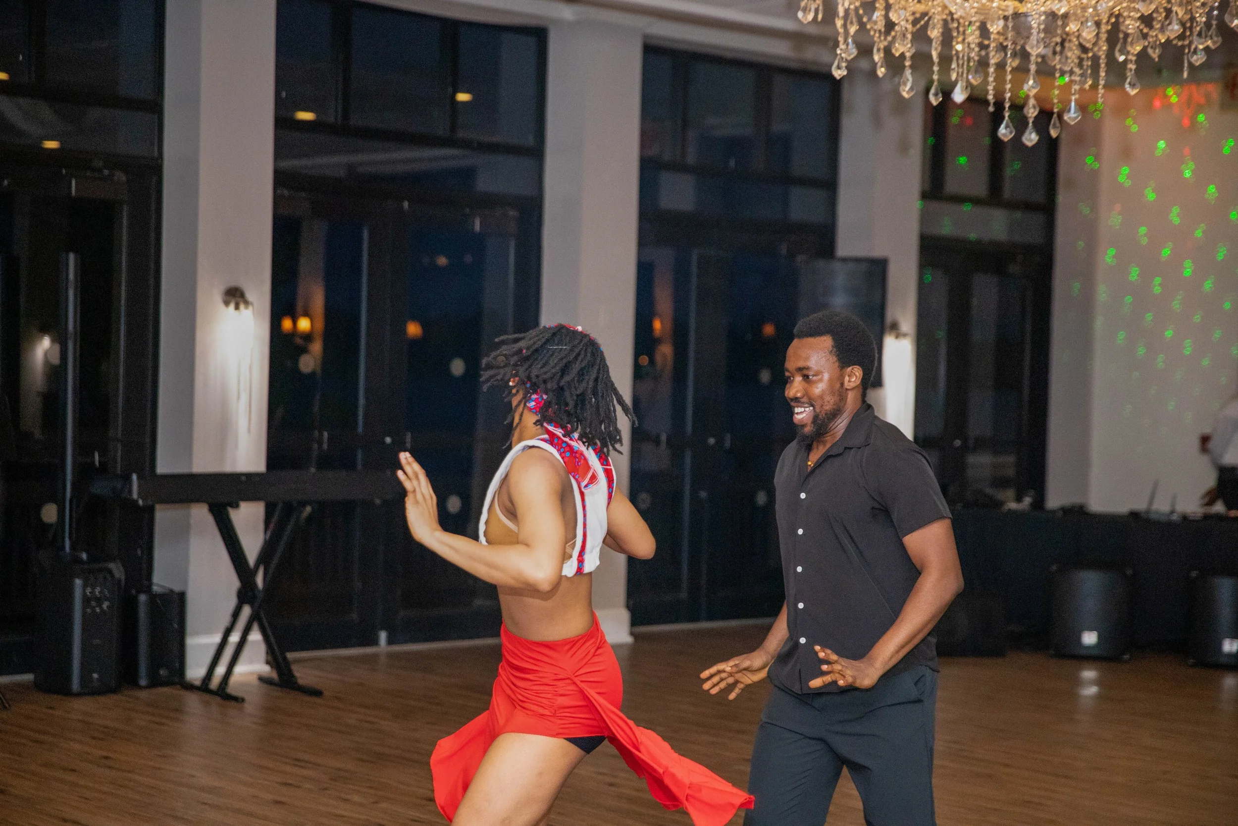 A man and woman dancing together indoors at a social event, with the woman wearing a red skirt and the man wearing dark pants and a shirt. They are smiling and looking at each other under a chandelier.