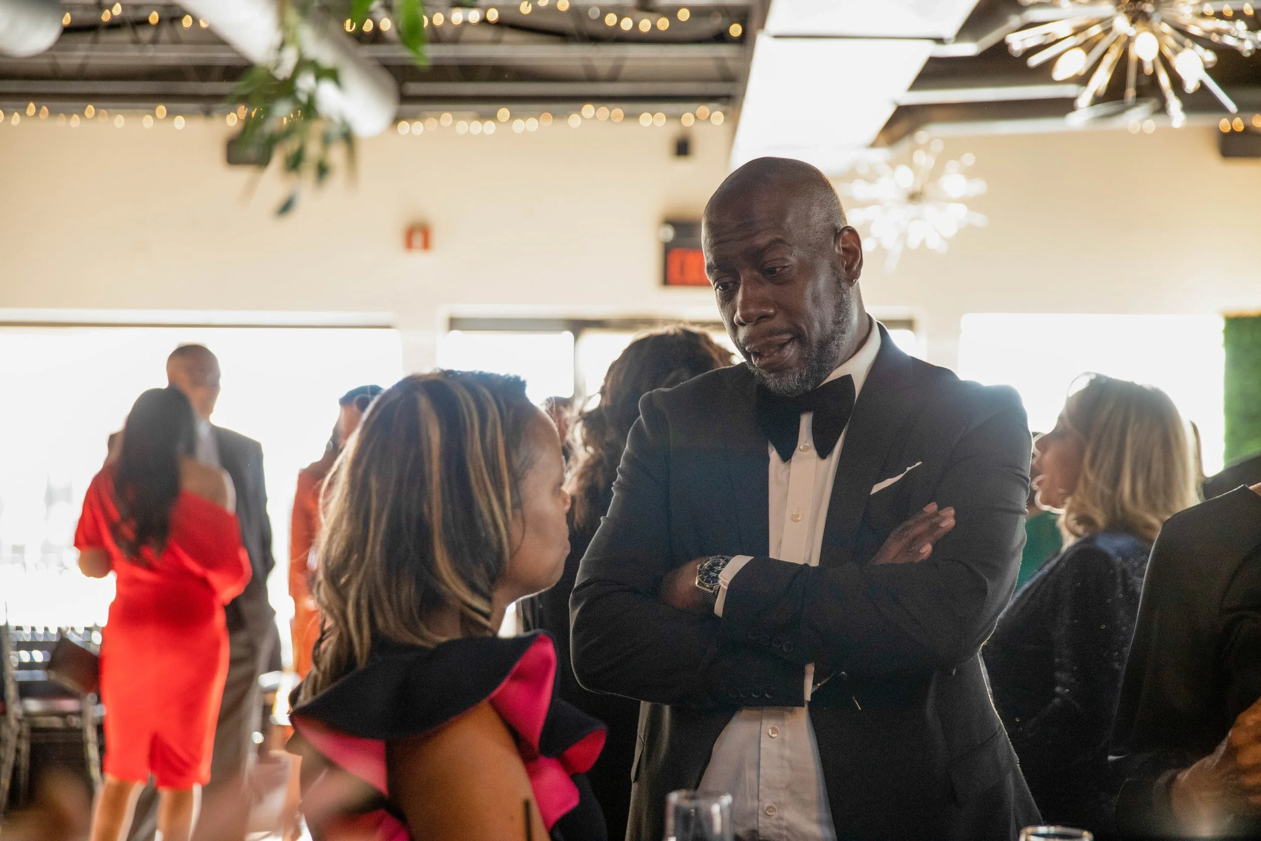 A man in a tuxedo with a bow tie is talking to a woman at a formal event. There are several other people engaged in conversation in the background. The setting appears to be a decorated banquet hall with string lights and chandeliers.
