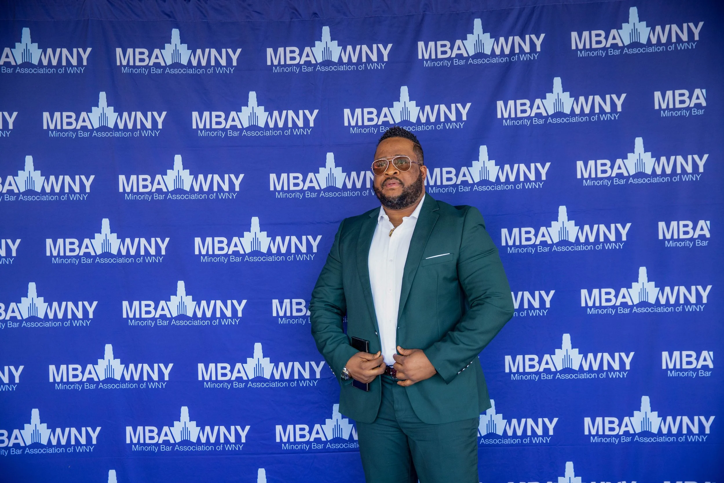 A man wearing sunglasses and a dark green suit standing in front of a blue backdrop with white text and logos for the Minority Bar Association of WNY.