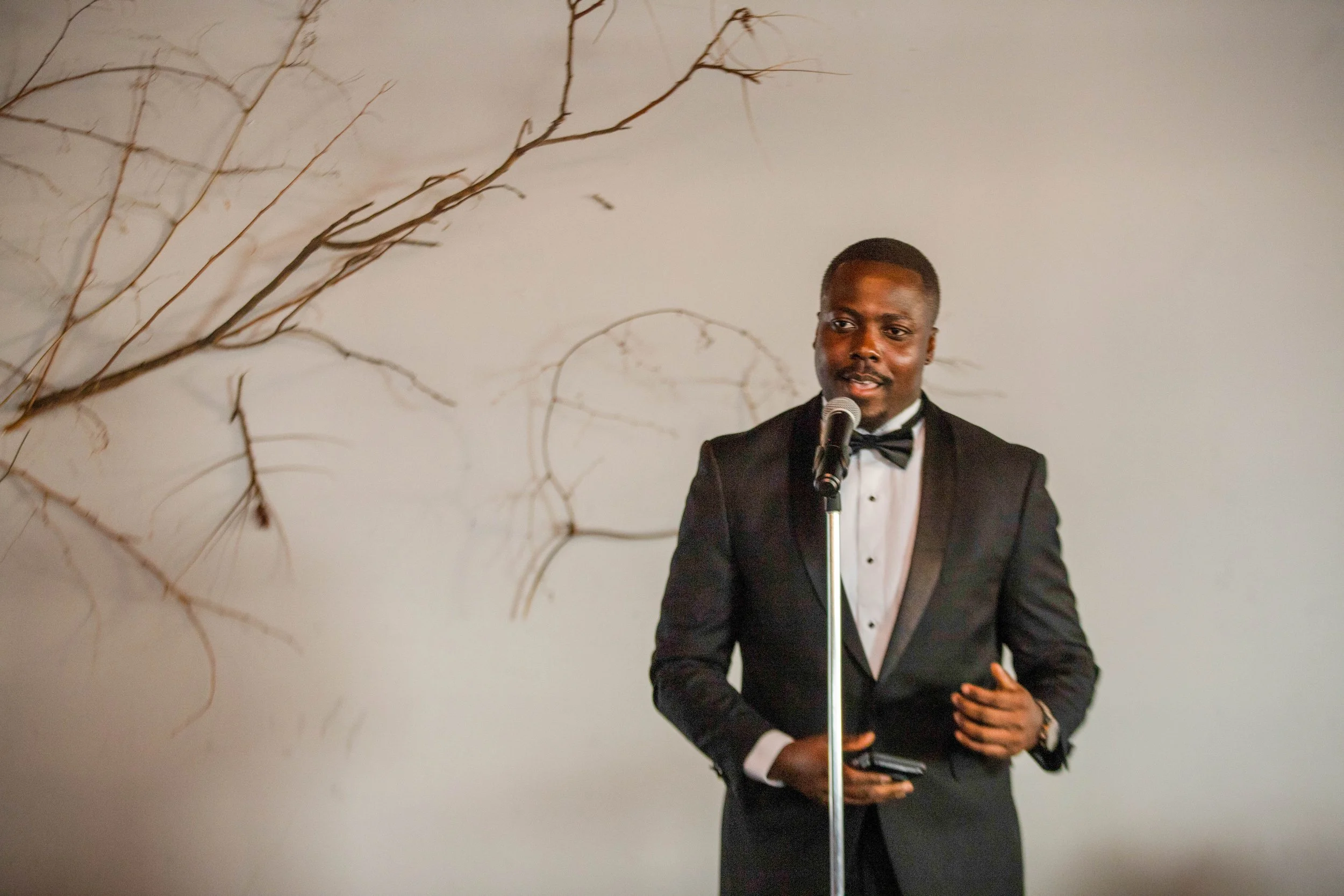 A man in a tuxedo speaking at a microphone in front of a plain wall with some artful branches.