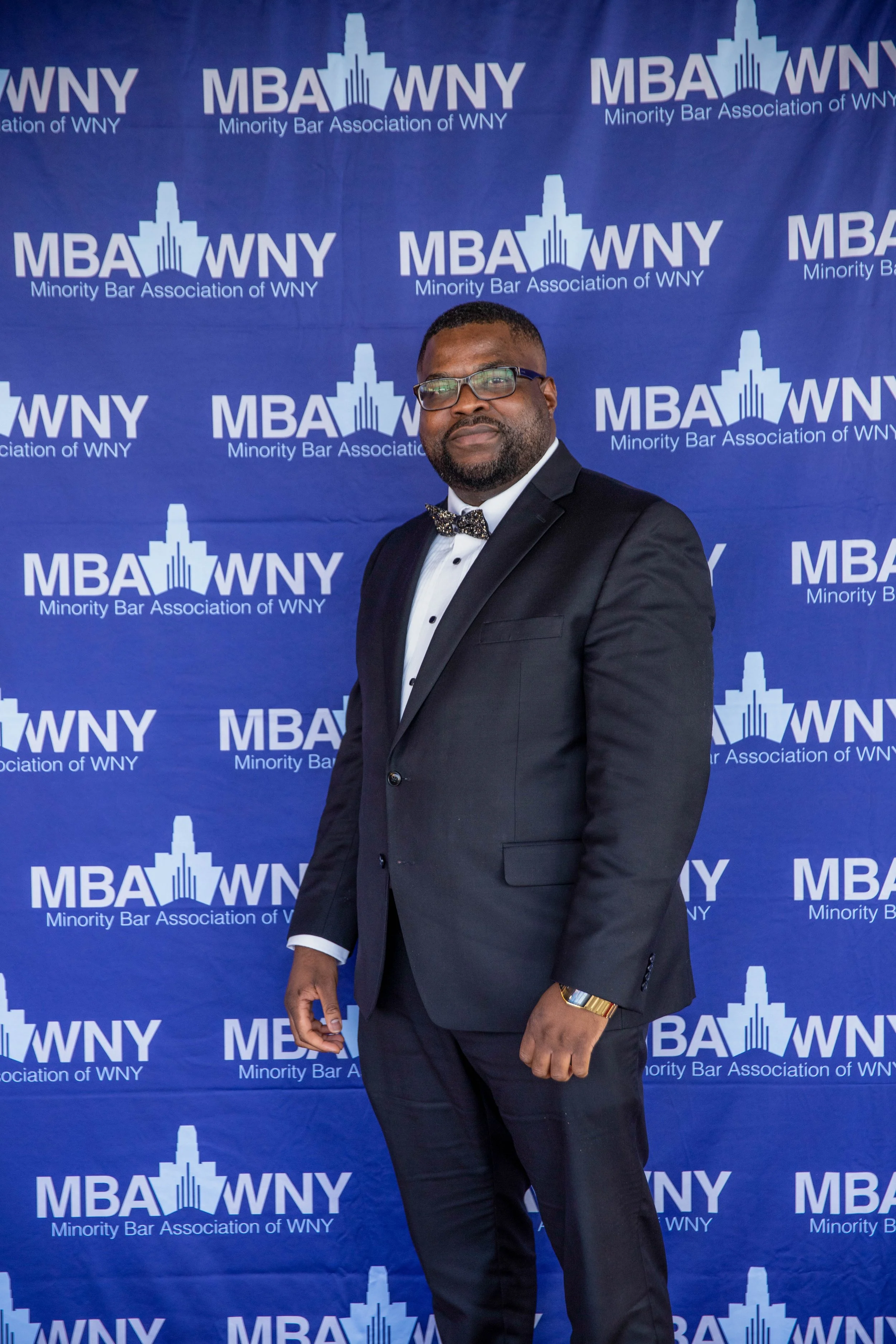 A man in a black tuxedo, white dress shirt, and bow tie standing in front of a blue backdrop with the text 'MBA WNY Minority Bar Association of WNY' repeatedly printed on it. He is wearing glasses, a gold watch, and has a beard.