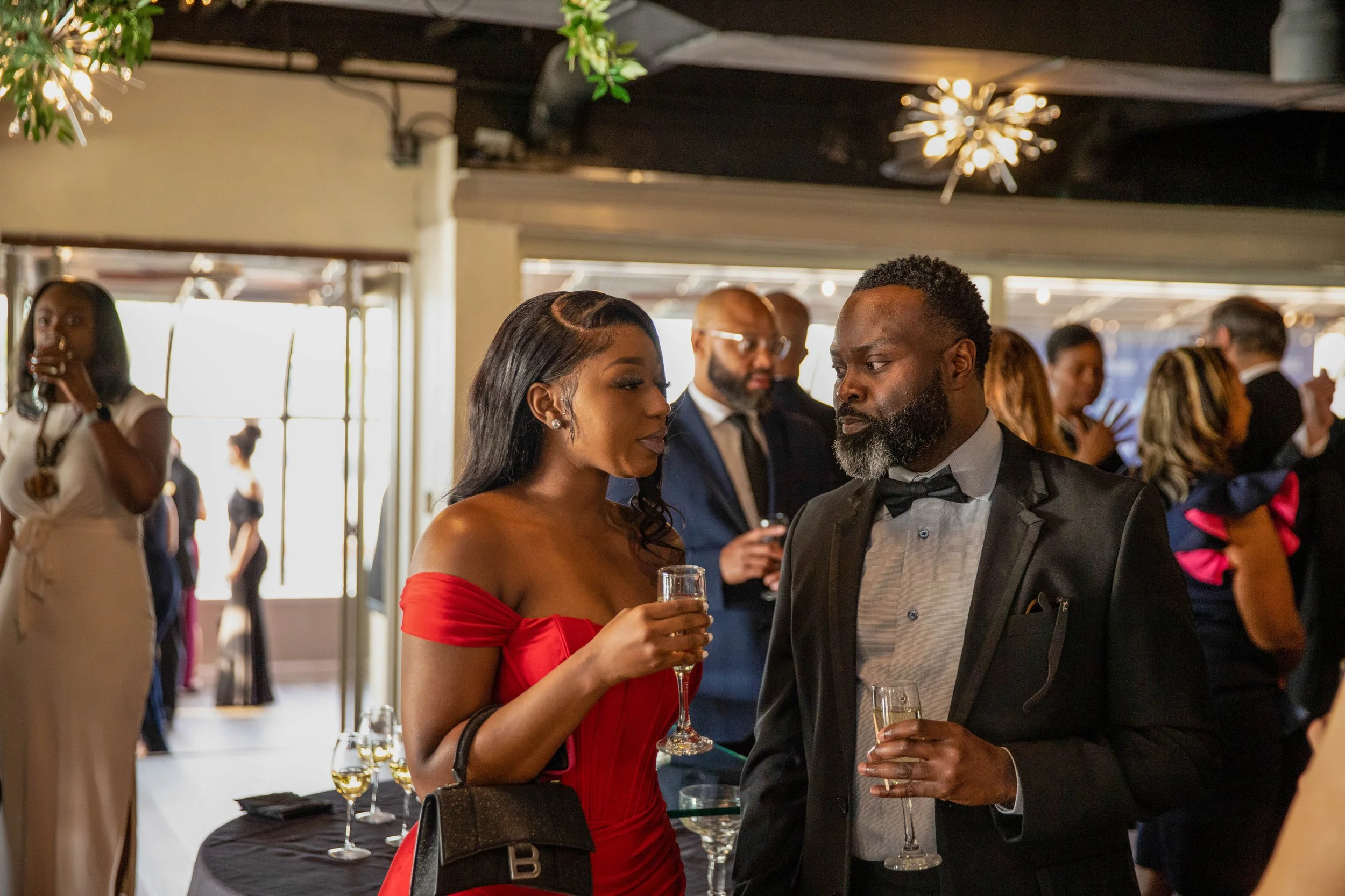 A woman in a red off-shoulder dress holding a glass of champagne and speaking with a man in a black tuxedo at a social event. The background shows other people engaging and a decorated indoor space with warm lighting.
