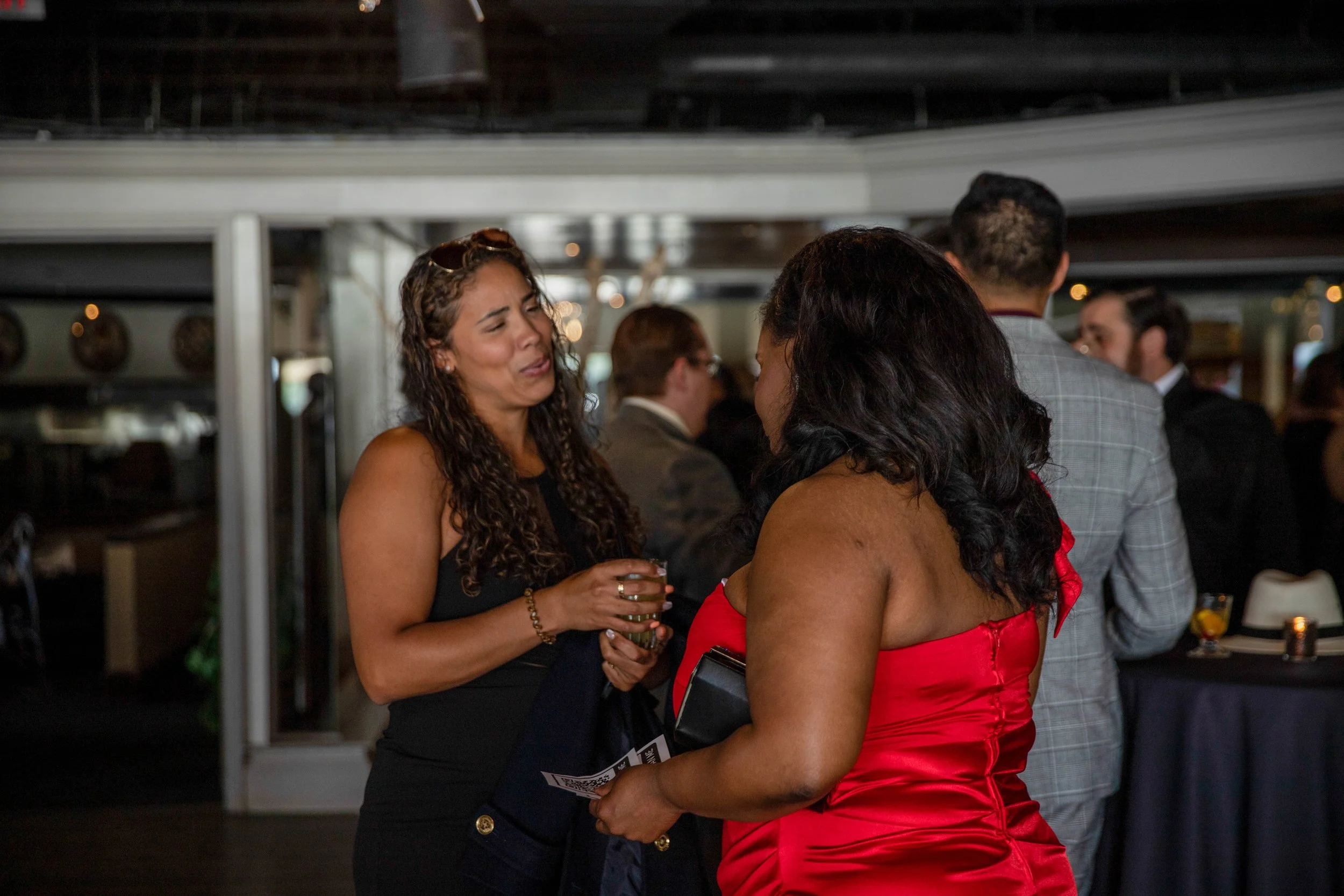 Two women are conversing at a social event. One woman, wearing a black dress and sunglasses on her head, is holding a drink. The other woman, dressed in a red satin gown, has dark, curly hair.