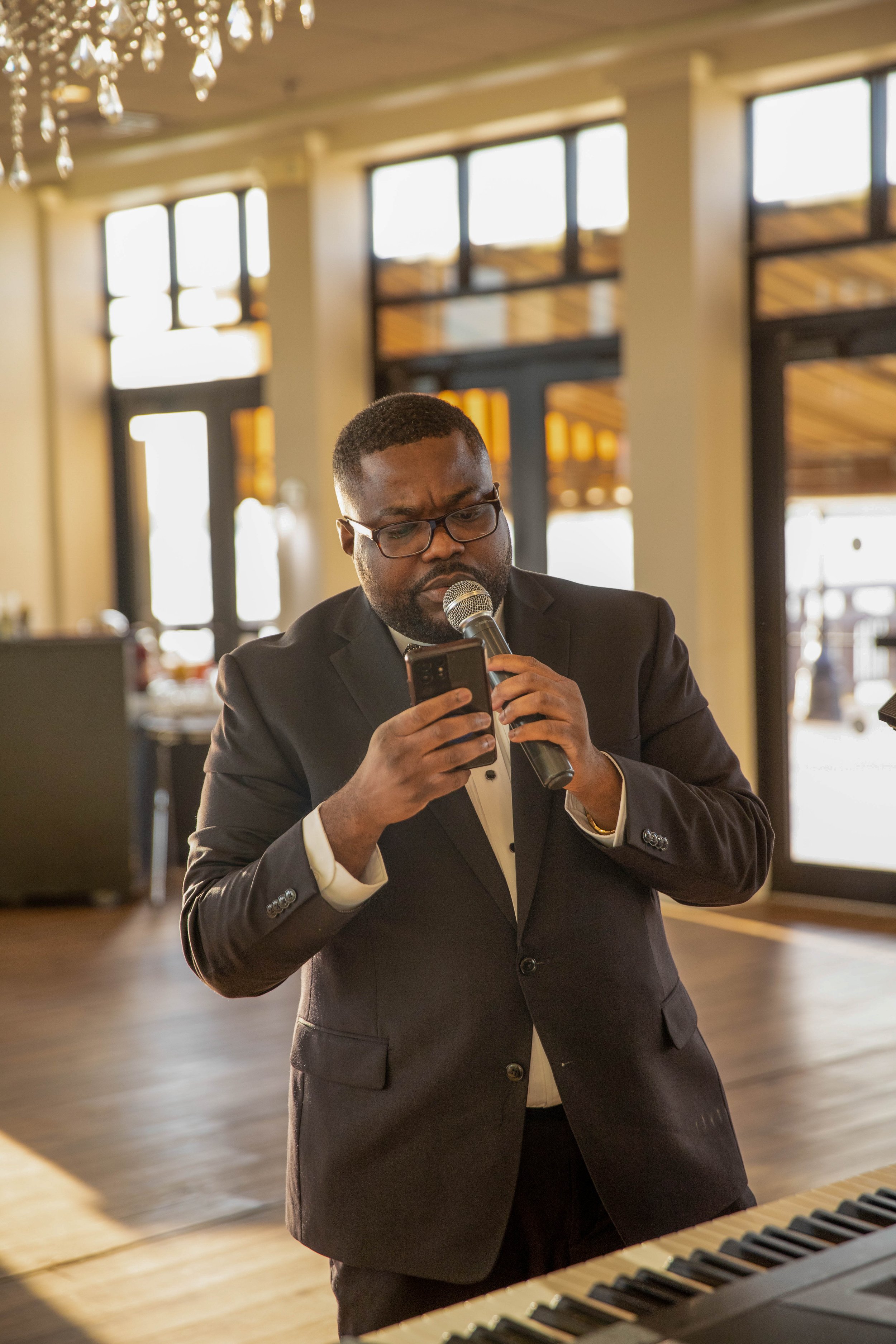 A man in a black suit wearing glasses, holding a microphone and looking at his phone, standing in a well-lit room with large windows and a piano in the foreground.
