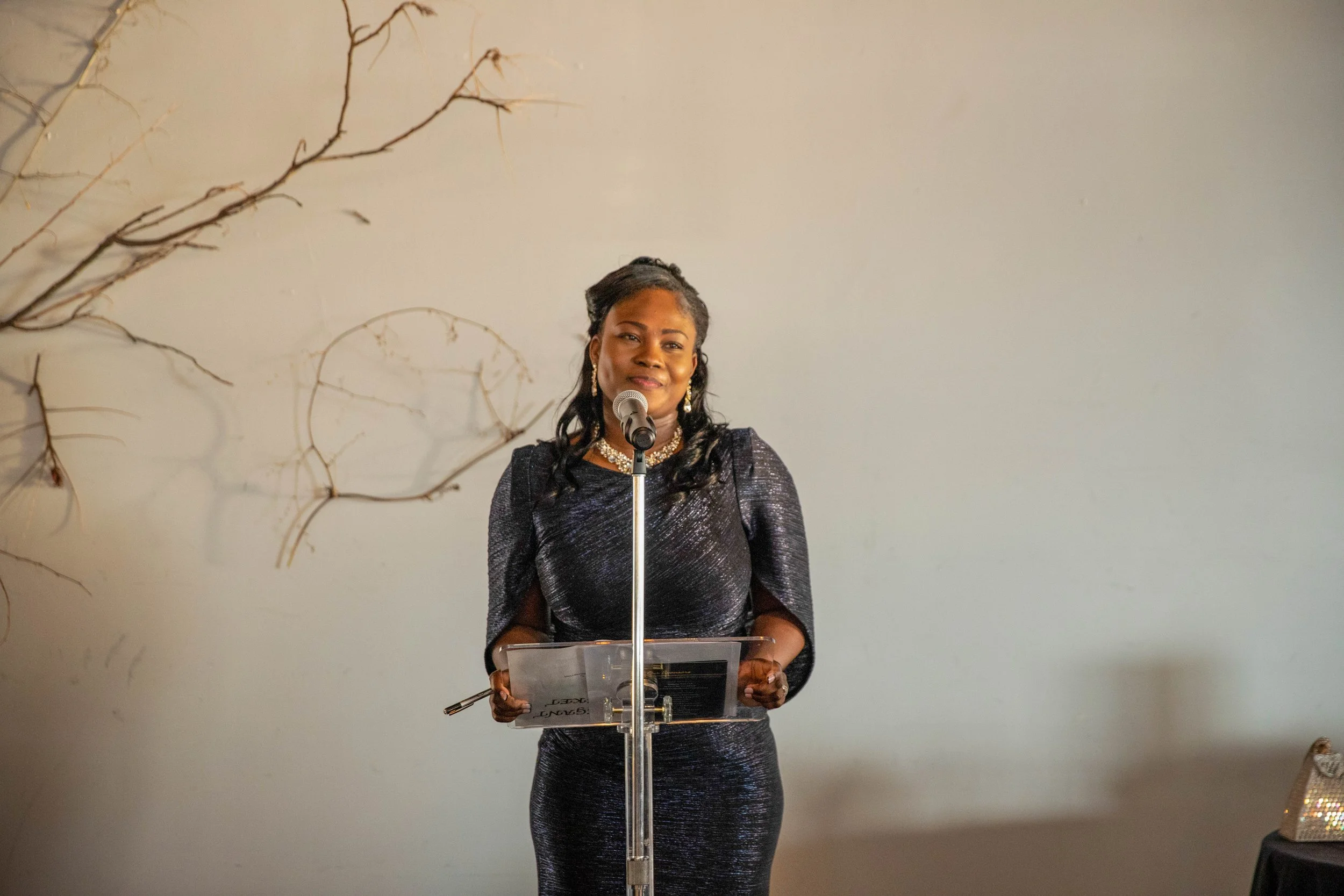 A woman in a black, shiny dress stands behind a clear podium, speaking into a microphone, with a neutral-colored wall behind her decorated with a few thin, bare tree branches.