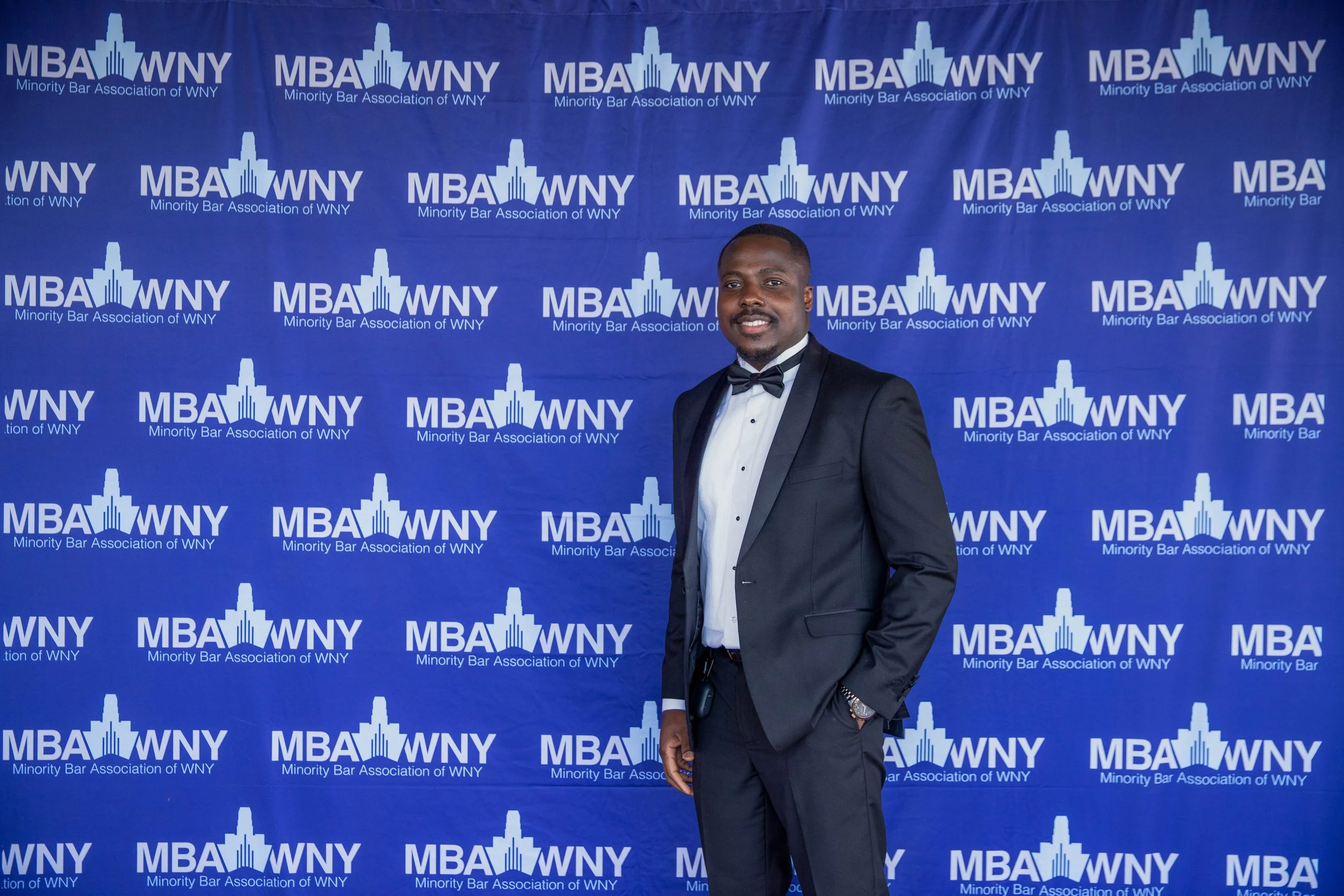 A man in a tuxedo and bow tie standing in front of a blue backdrop with the logo and text 'MBA WNY Minority Bar Association of WNY' repeated across it.