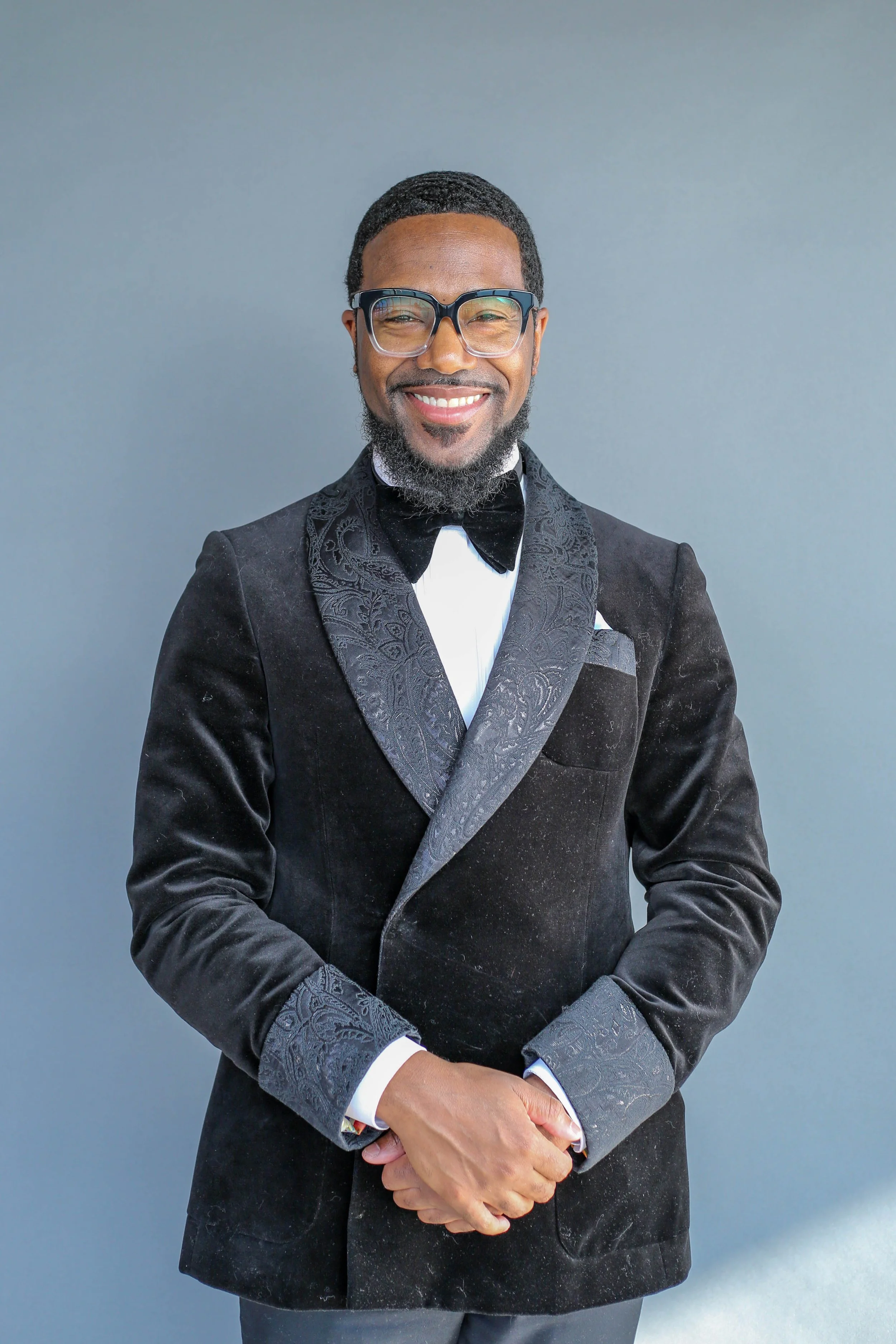 A man in a black tuxedo with a patterned lapel and cuffs, wearing large black-framed glasses, smiling with white teeth, with a well-groomed beard, standing against a gray background.