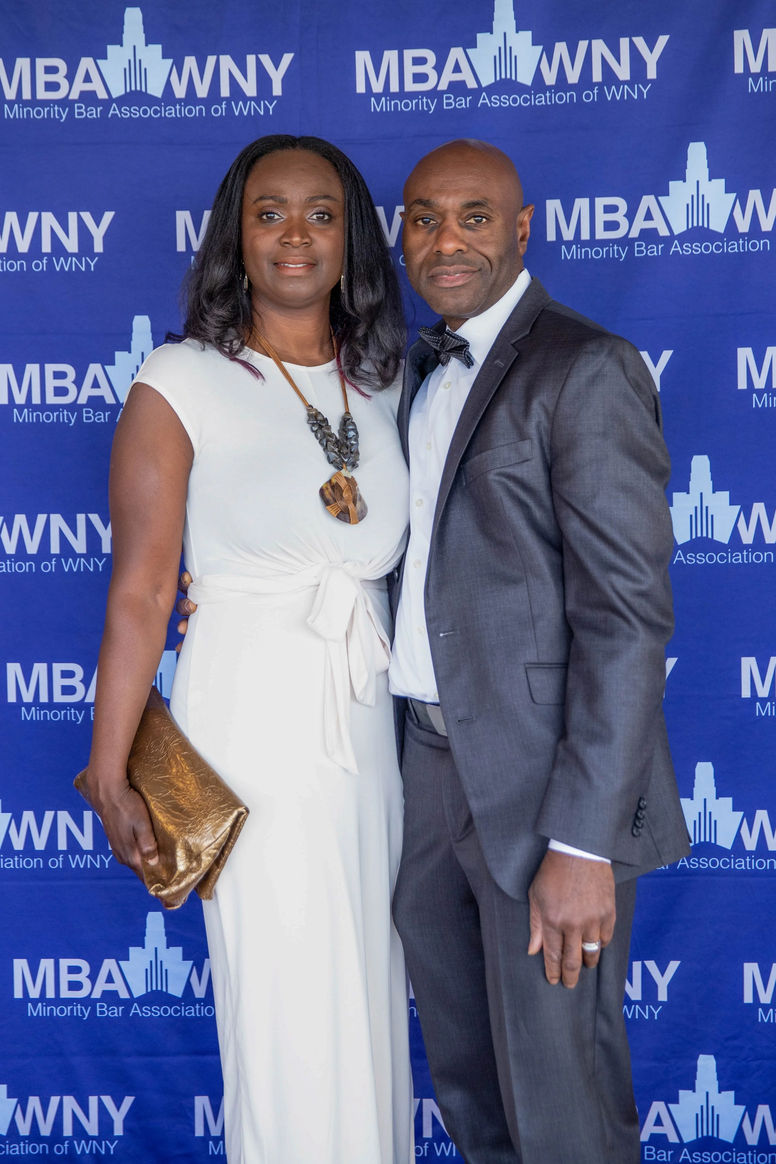 A man and woman dressed in formal attire posing in front of a backdrop with the logo 'MBA WNY' and text 'Minority Bar Association of WNY'. The woman is wearing a white dress with a statement necklace and holding a brown clutch, the man is in a gray s