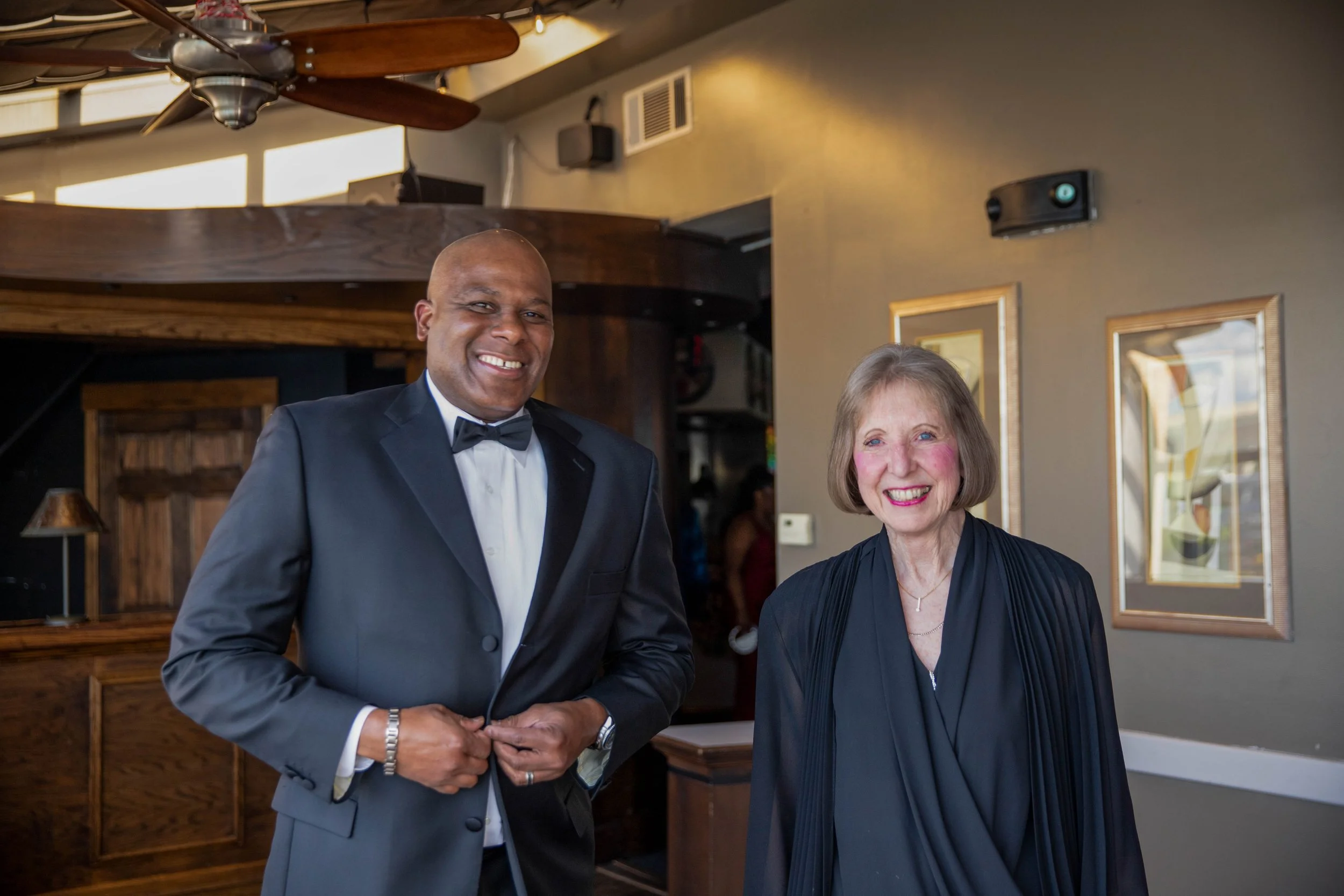 A man in a tuxedo and a woman in a black outfit smiling and shaking hands inside a restaurant or event space.