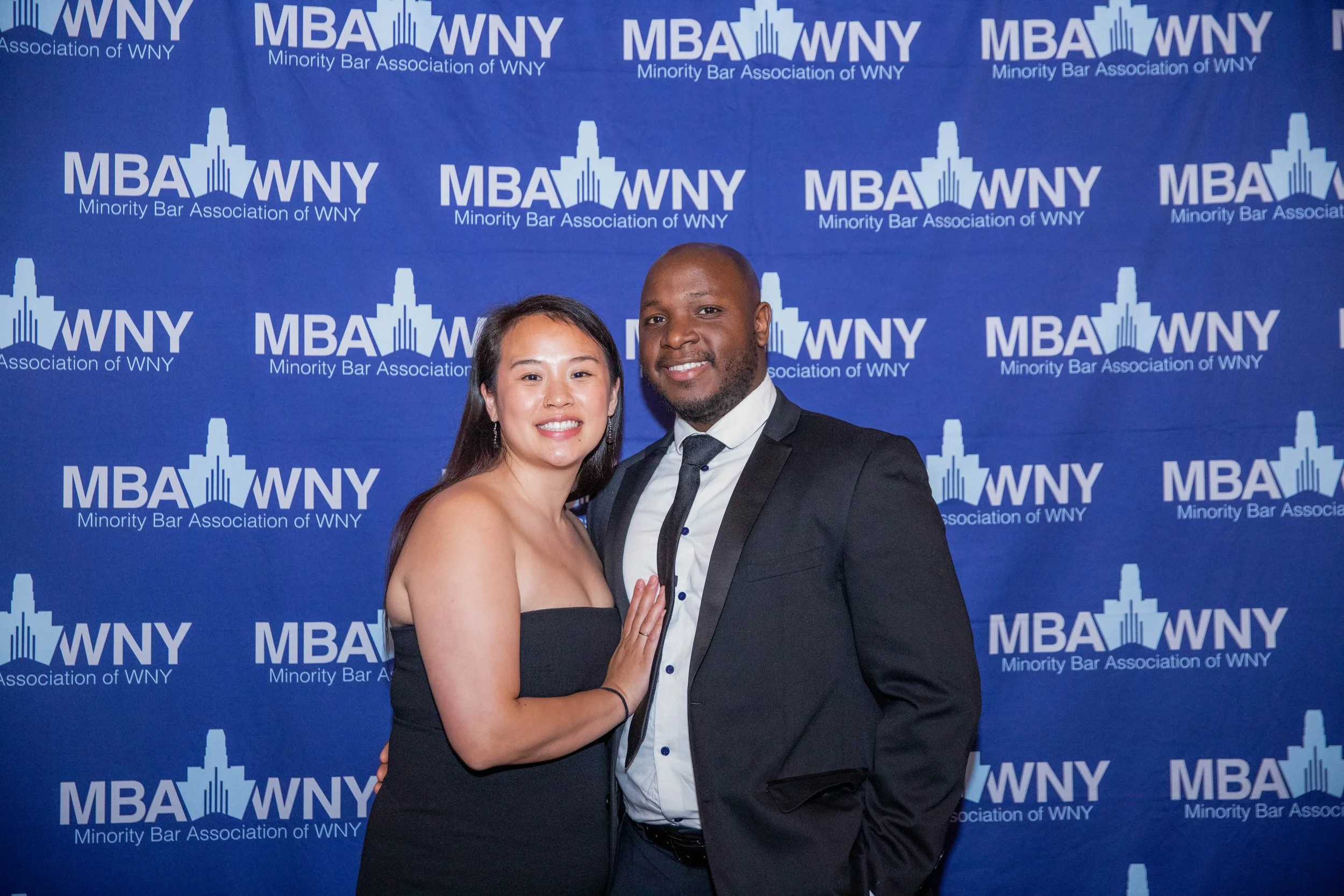 A man and woman are standing close together, smiling, at an event with a backdrop that reads 'Minority Bar Association of WNY' and 'MBA WNY.'