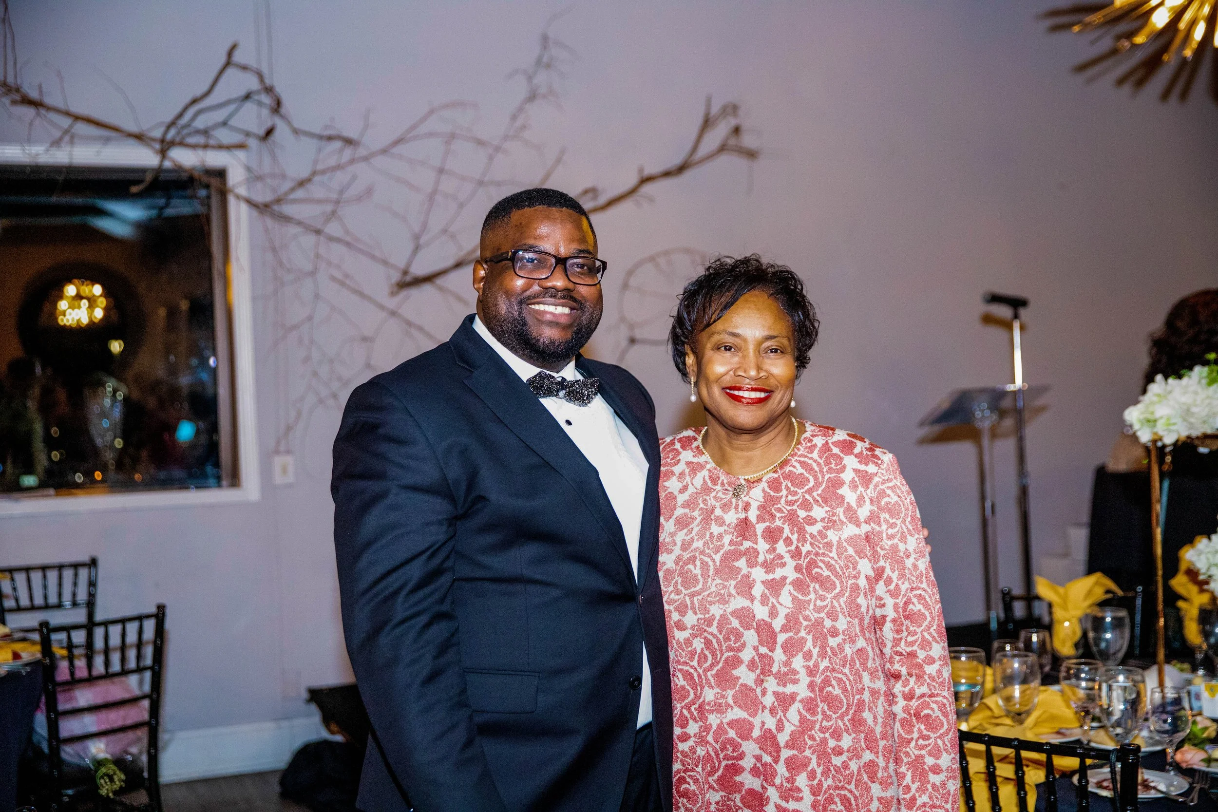 A smiling man in a tuxedo with a bowtie and glasses standing next to a smiling woman in a pink patterned dress at a formal event. There are tables with decorated place settings and a mirror reflecting the room's interior.