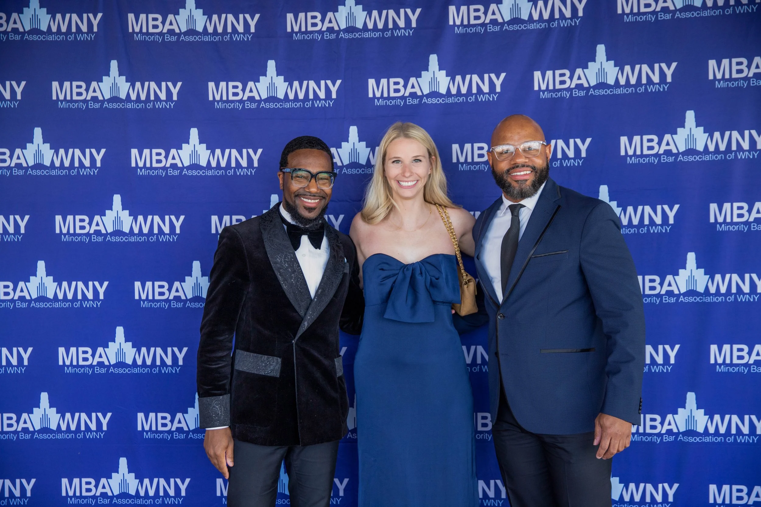 Three people standing in front of a blue backdrop with the logo ‘MBA WNY’ and text ‘Minority Bar Association of WNY’. The group includes two men and one woman, dressed in formal attire, smiling for the photo.
