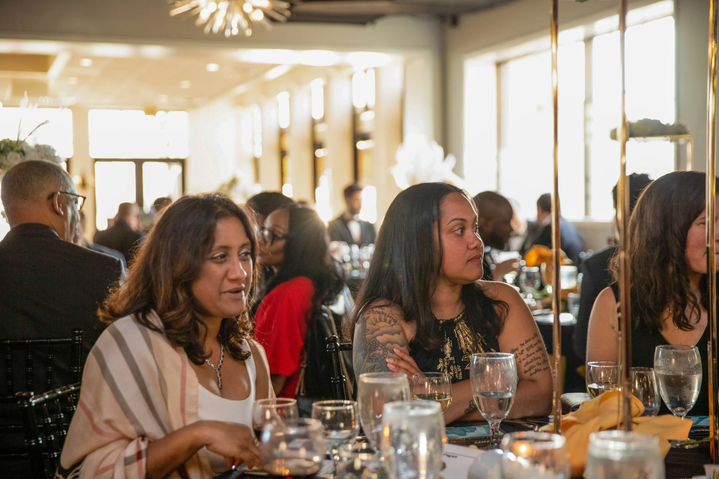 People sitting at a formal event dining table with glasses of water and wine, some women with tattoos, in a well-lit room with large windows, gold accents, and multiple guests in the background.