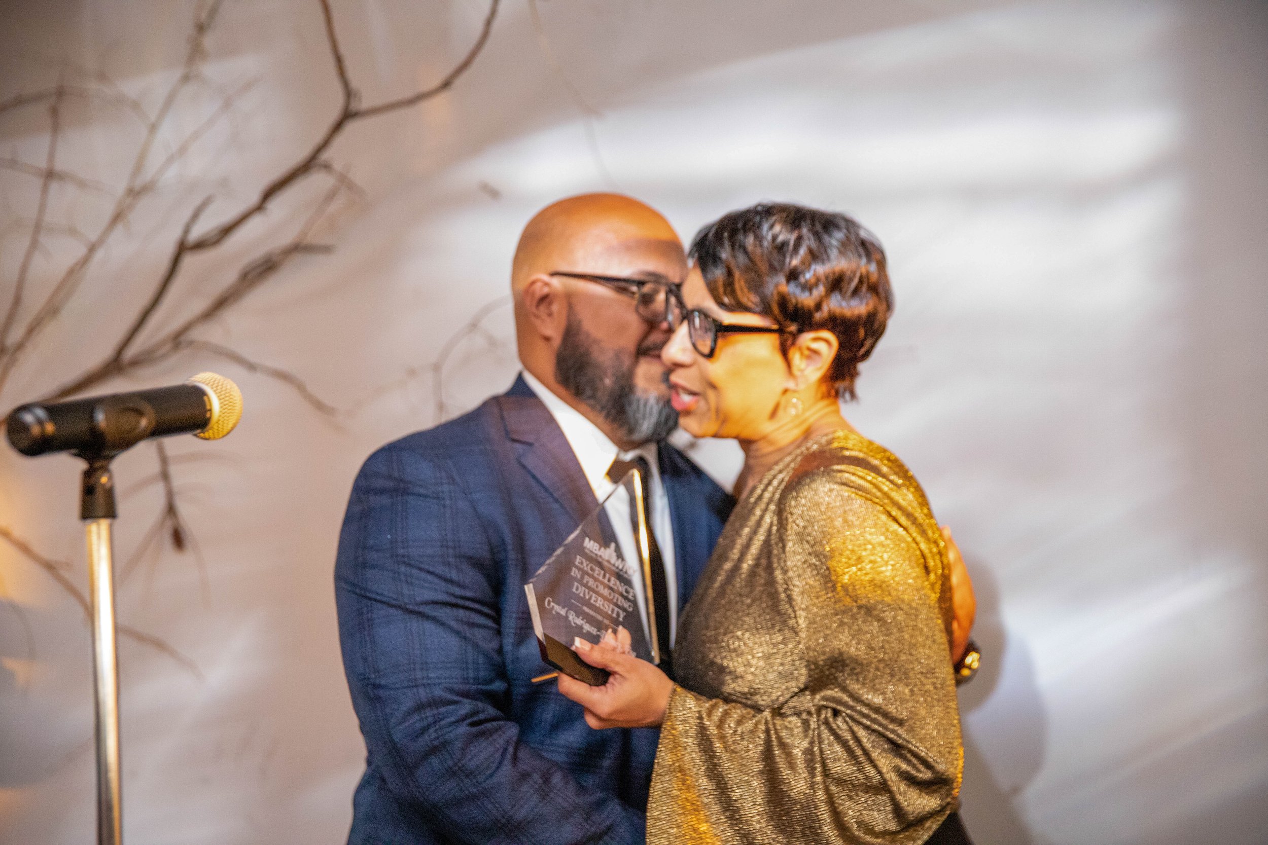A man and woman face each other, close up, during a handshake or embrace at an event. The man is bald with a beard, wearing glasses and a blue suit. The woman has short dark hair, styled with waves, and is wearing glasses and a shiny gold outfit. The