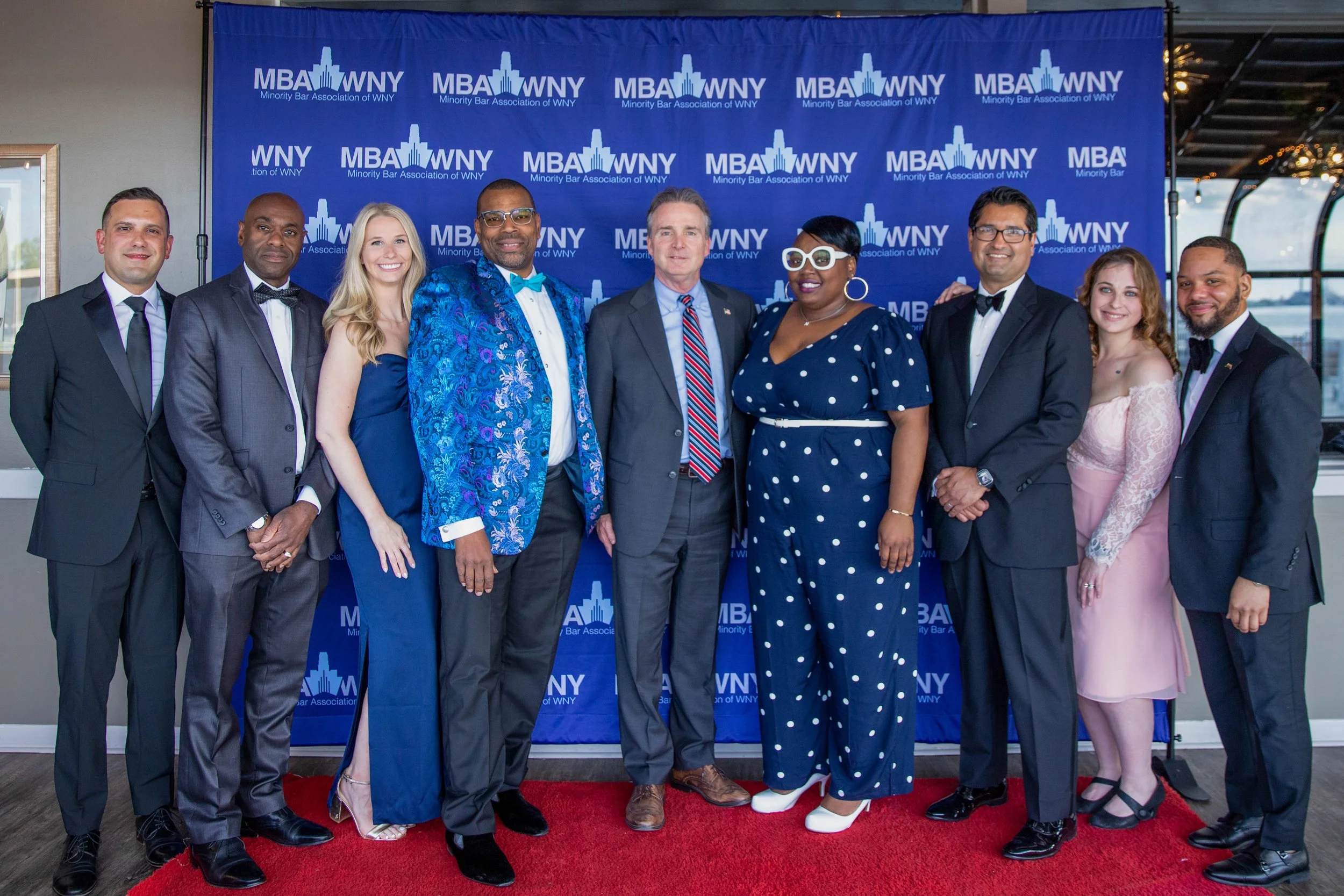 Group of nine diverse people standing on a red carpet in front of a blue backdrop with the logo of the Minority Bar Association of WNY. They are dressed in formal attire, smiling for the photo at a professional or celebratory event.