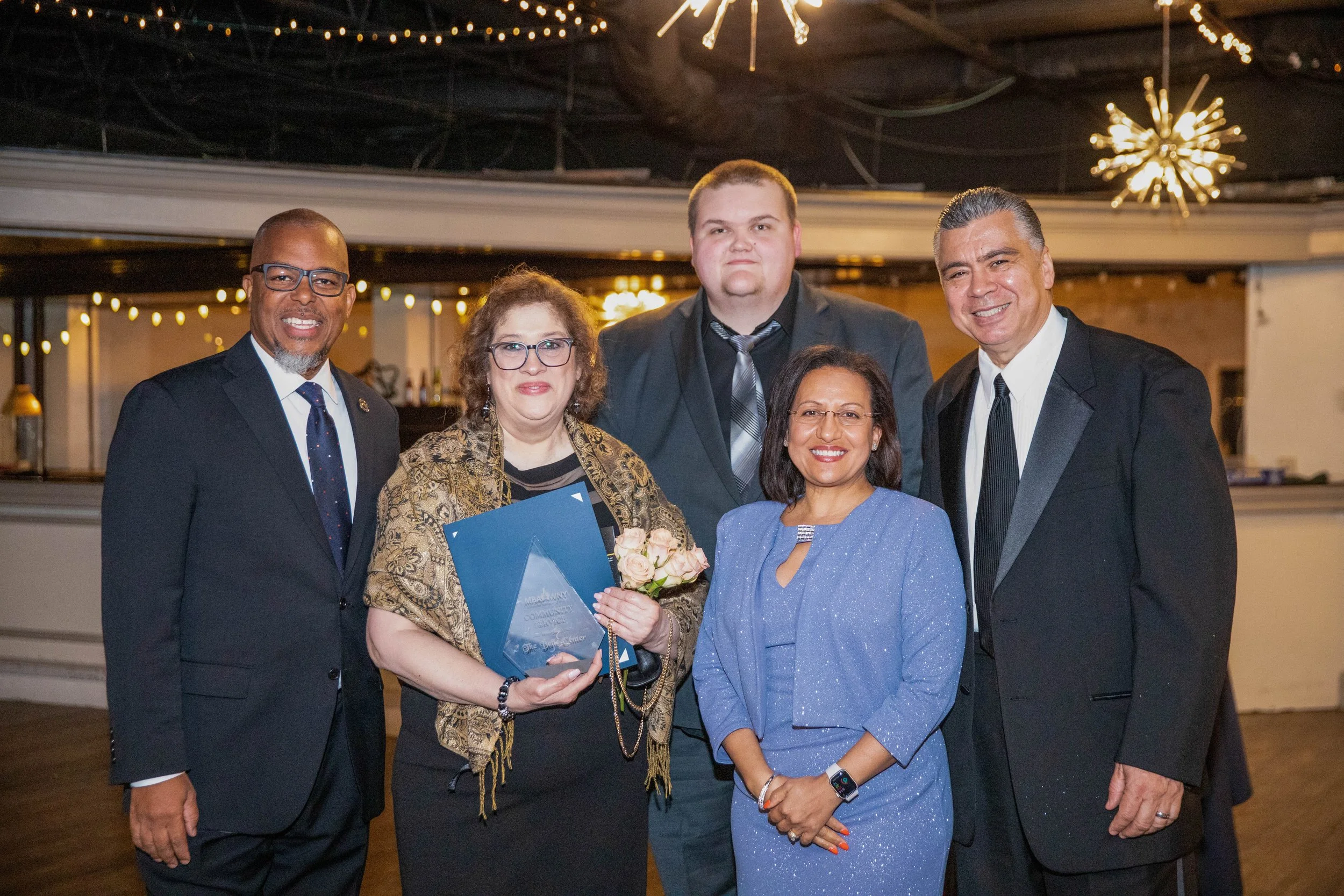 Group of six professionally dressed individuals gathered at an indoor event with warm lighting and decorative fixtures, posing for a photo, one woman holding a bouquet of flowers and awards.