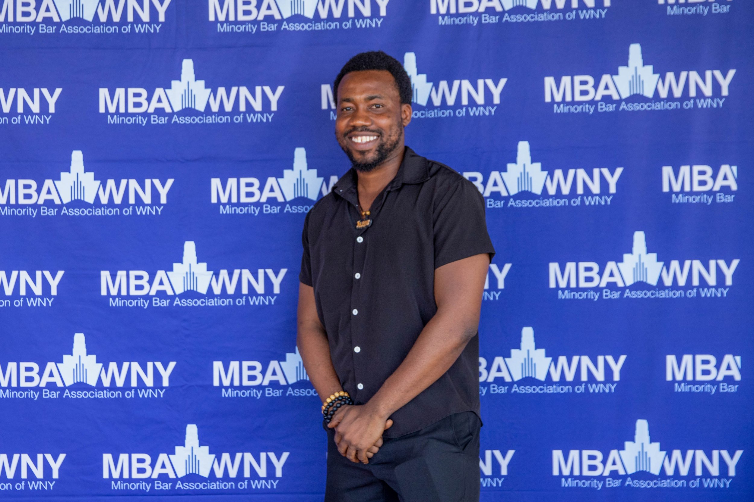A man in a black shirt smiling and standing in front of a blue backdrop with the white text and logo for the Minority Bar Association of WNY, including the letters MBA WNY and an abstract building logo.