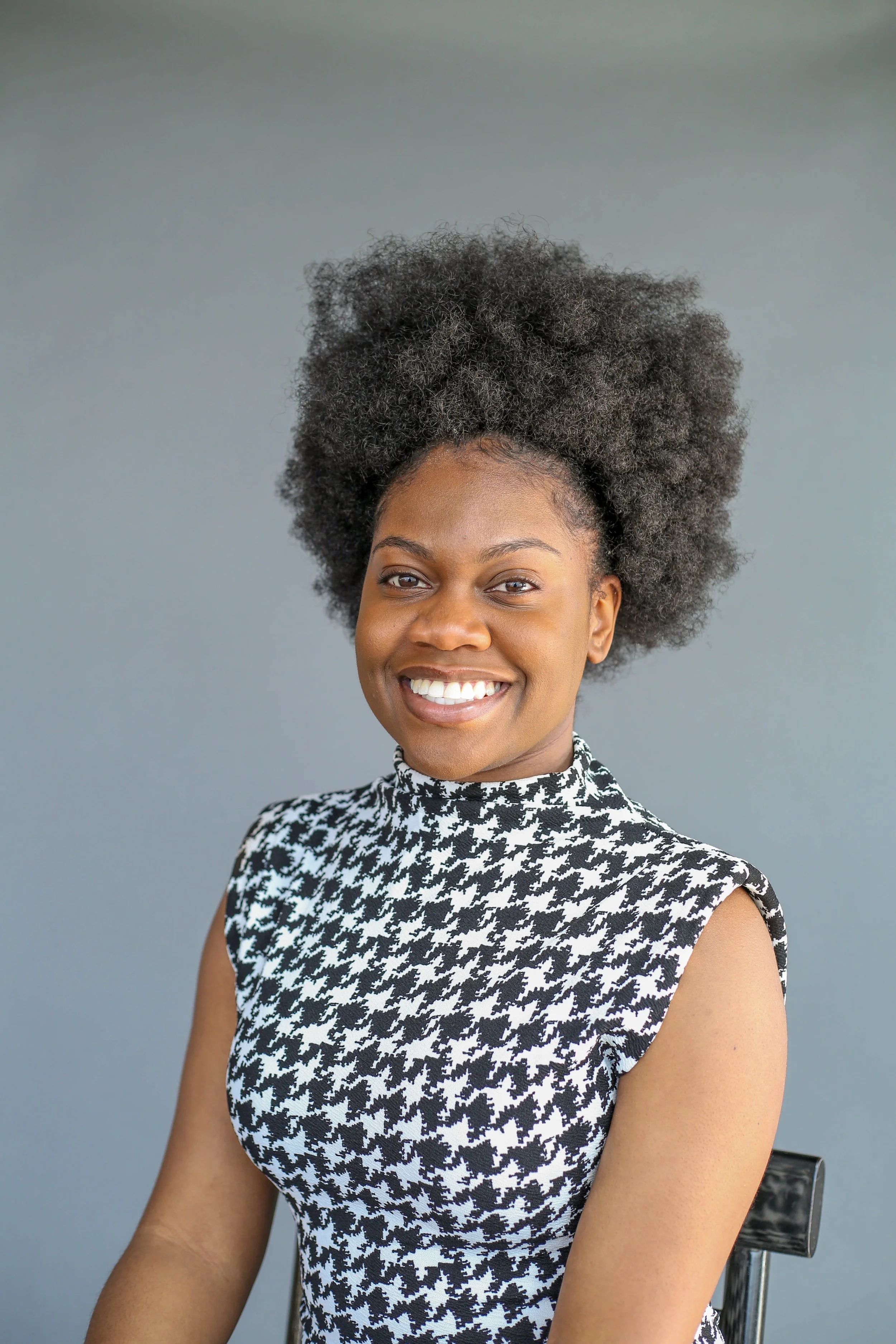 A smiling woman with dark skin and afro hair wearing a black and white houndstooth patterned sleeveless dress, sitting on a clear chair against a plain gray background.