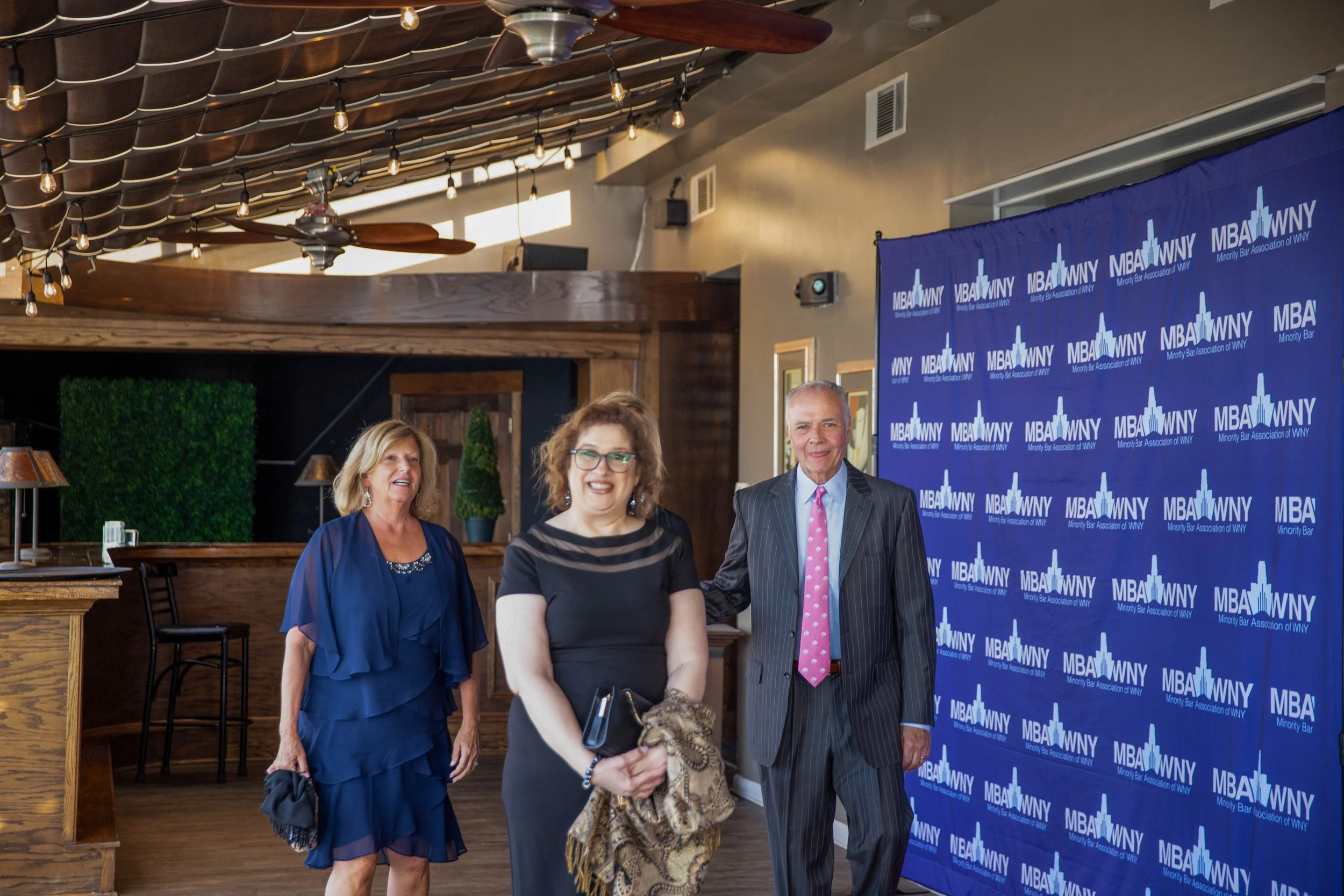 Three women and one man standing in front of a blue backdrop with the logo of MBA of WNY, at an indoor event. The man is wearing a suit with a pink tie, and the women are in business attire. The venue has wooden walls and ceiling fans.