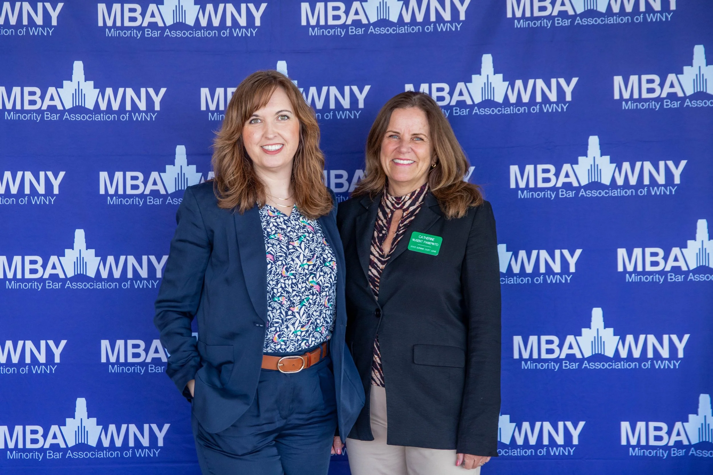 Two women standing in front of a blue backdrop with 'MBA WNY' and 'Minority Bar Association of WNY' printed on it, smiling at the camera.