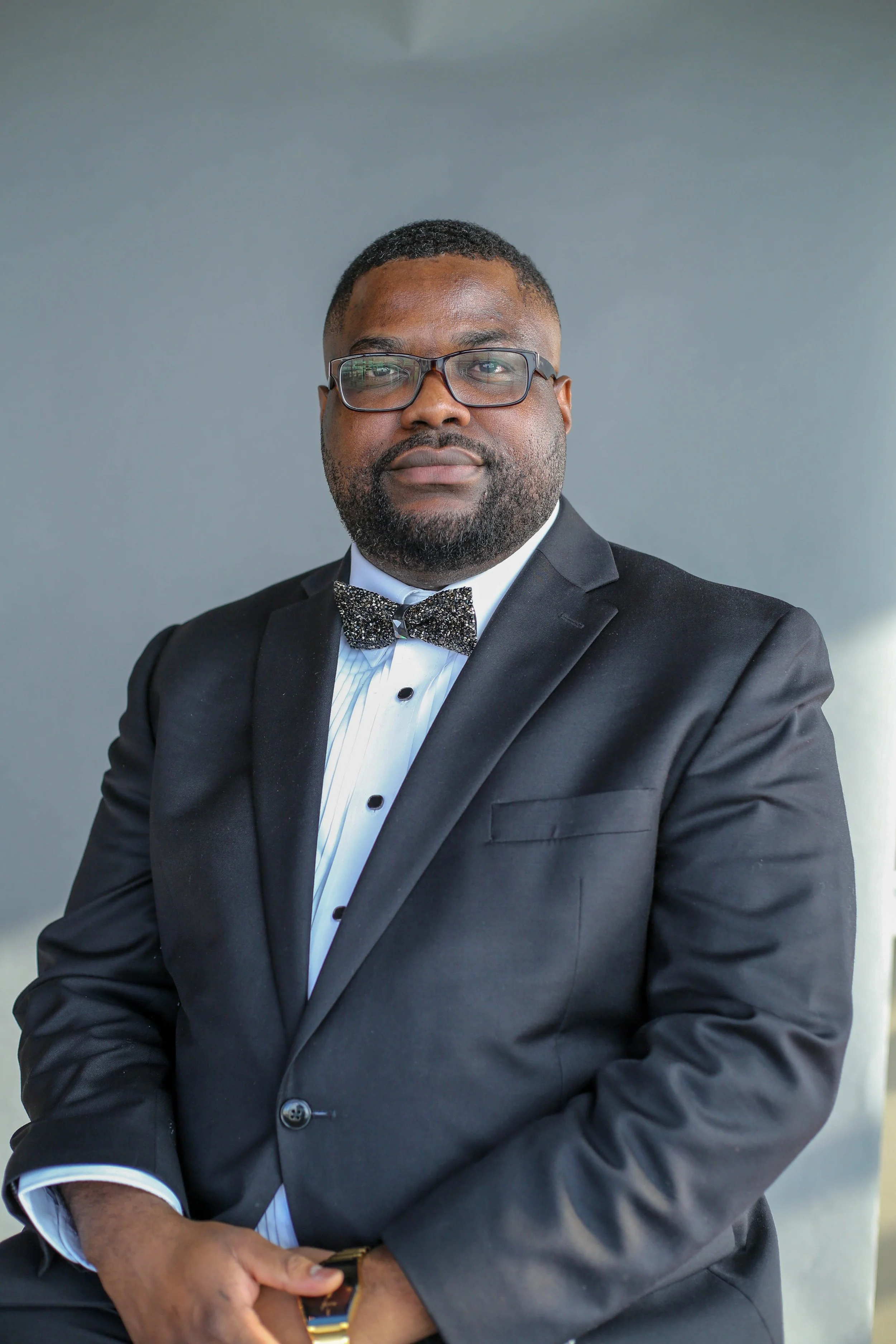 A man in a black tuxedo with a bowtie and glasses, posing with arms crossed against a plain background.