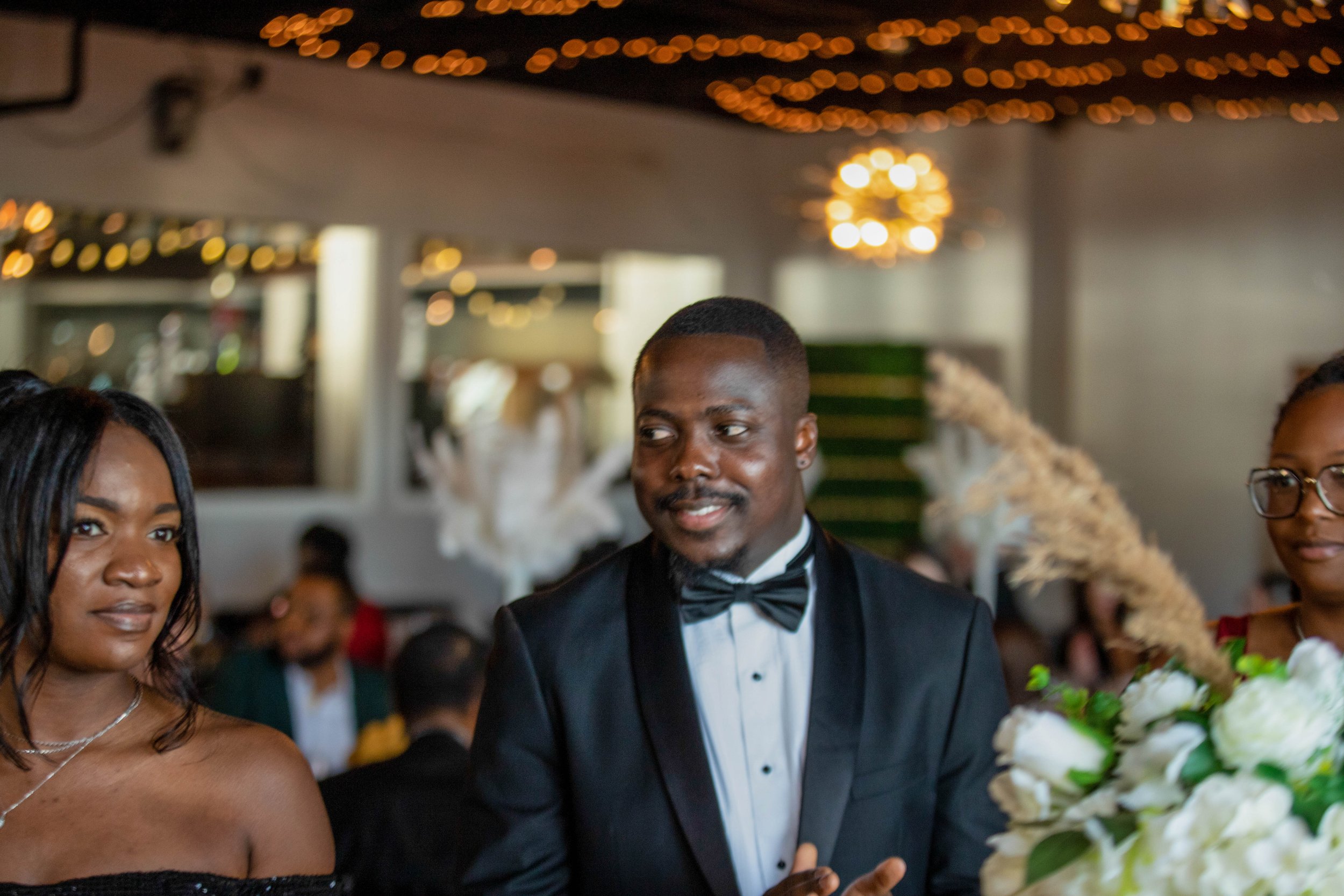 A man in a black tuxedo with a bow tie, smiling, standing next to a woman in a black dress at a wedding reception, with decorative ceiling lights and floral arrangements in the background.