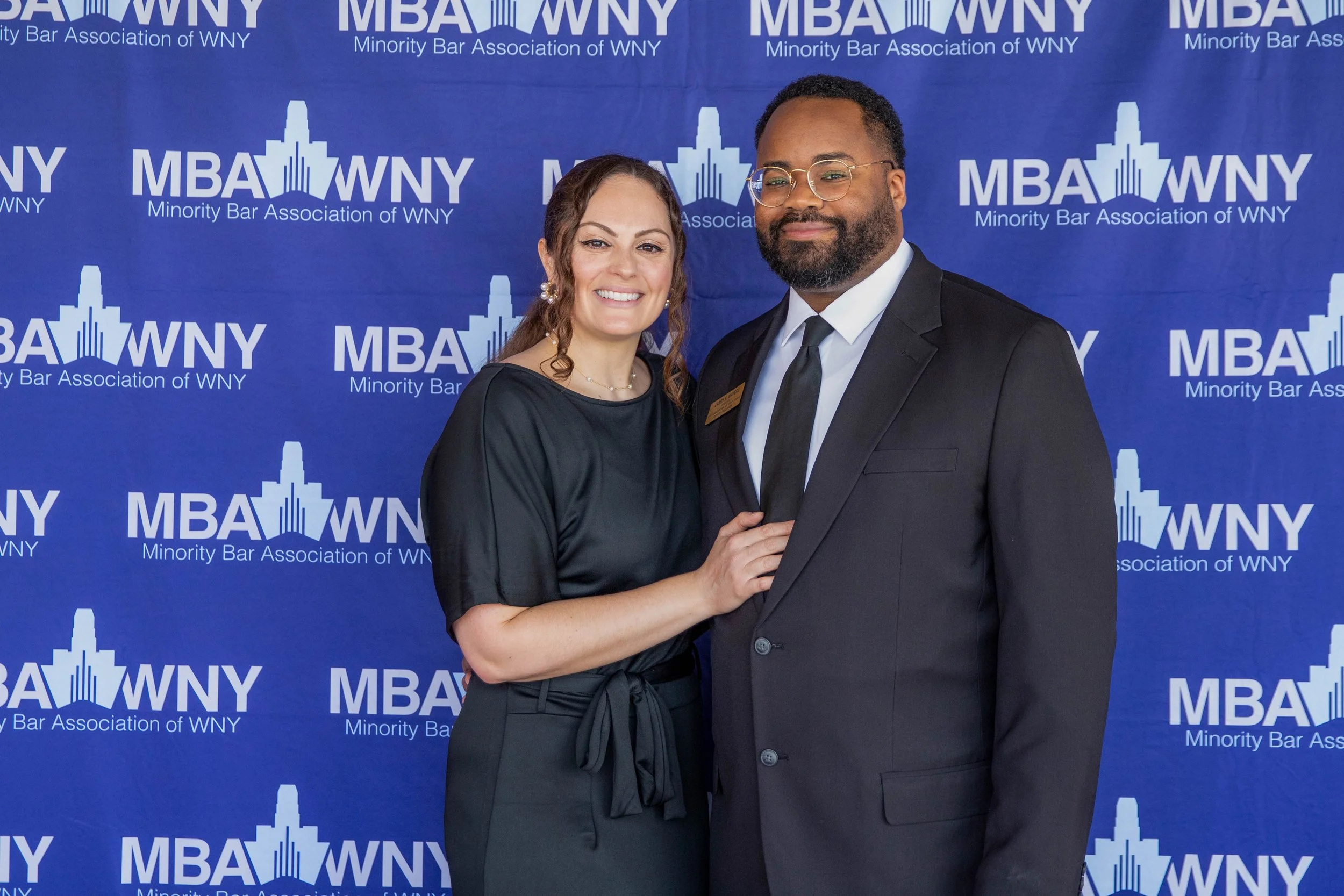 A man and a woman standing side by side smiling at the camera, dressed in formal attire with a blue backdrop featuring the logo and name of the Minority Bar Association of WNY.