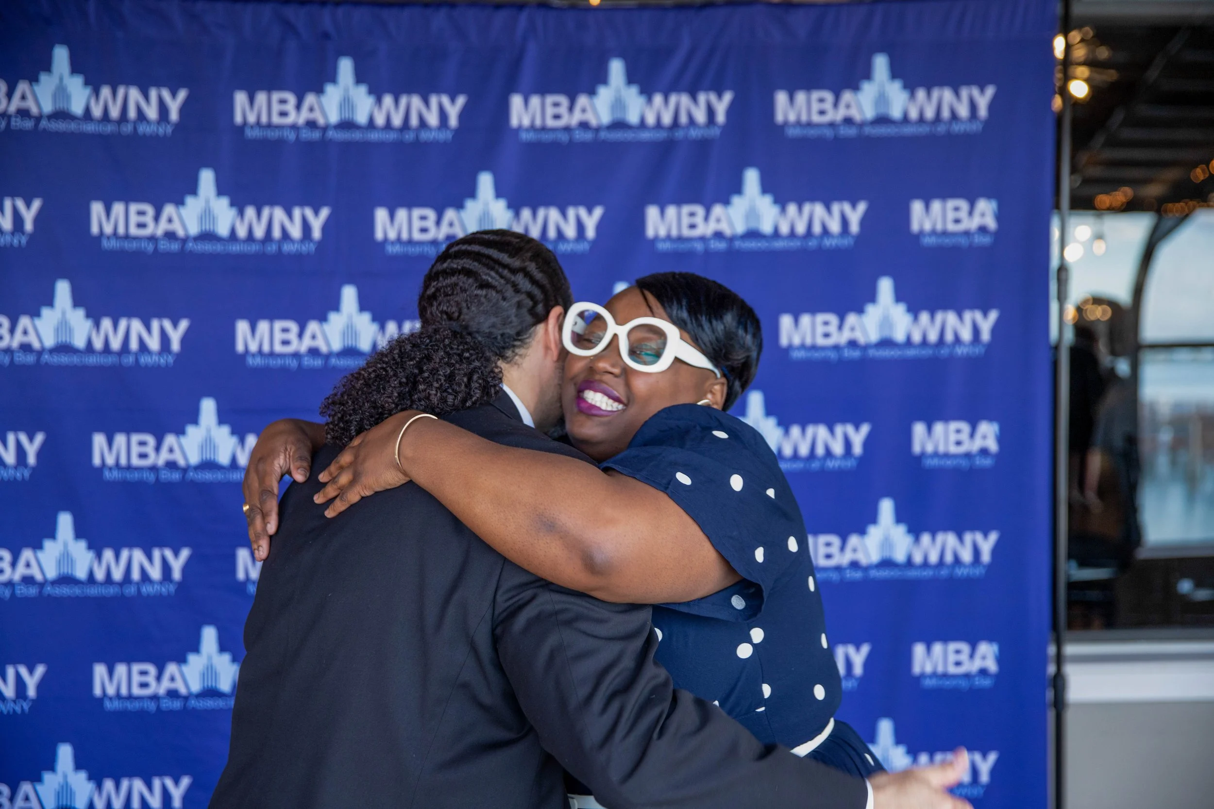 Two women hugging at a graduation event, with a blue backdrop featuring the MBA WNY logo.